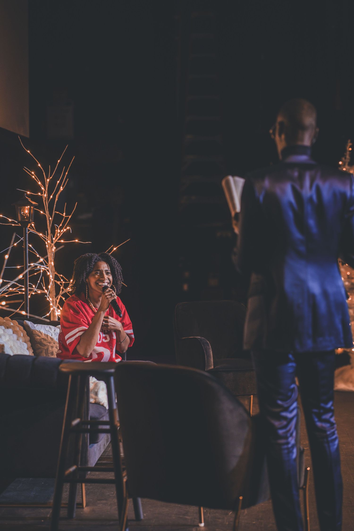 Stage performance with decorative lit branches and performers in conversation.