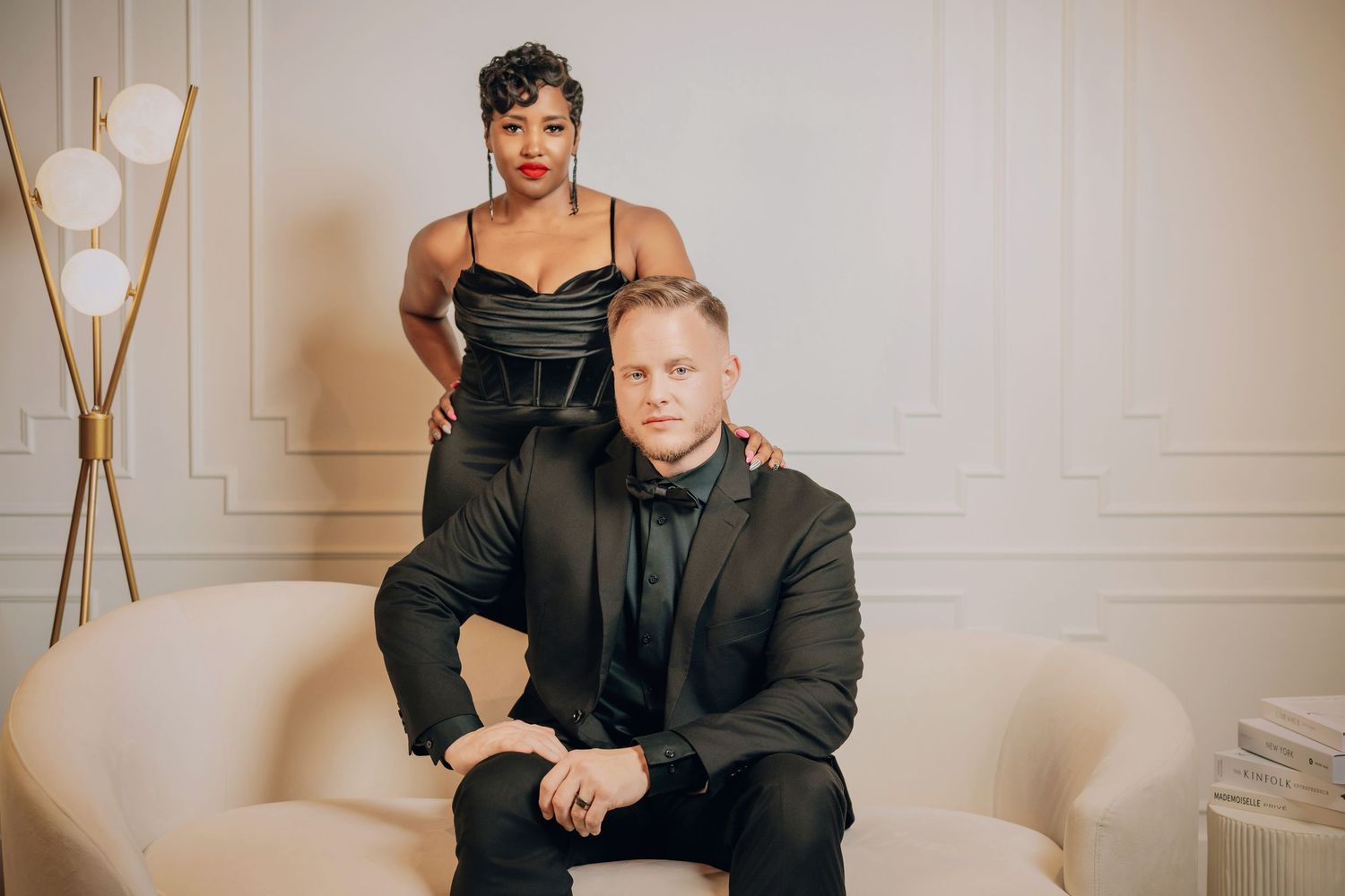 Two people pose together on a cream-colored couch in formal black attire with modern decor in the background.