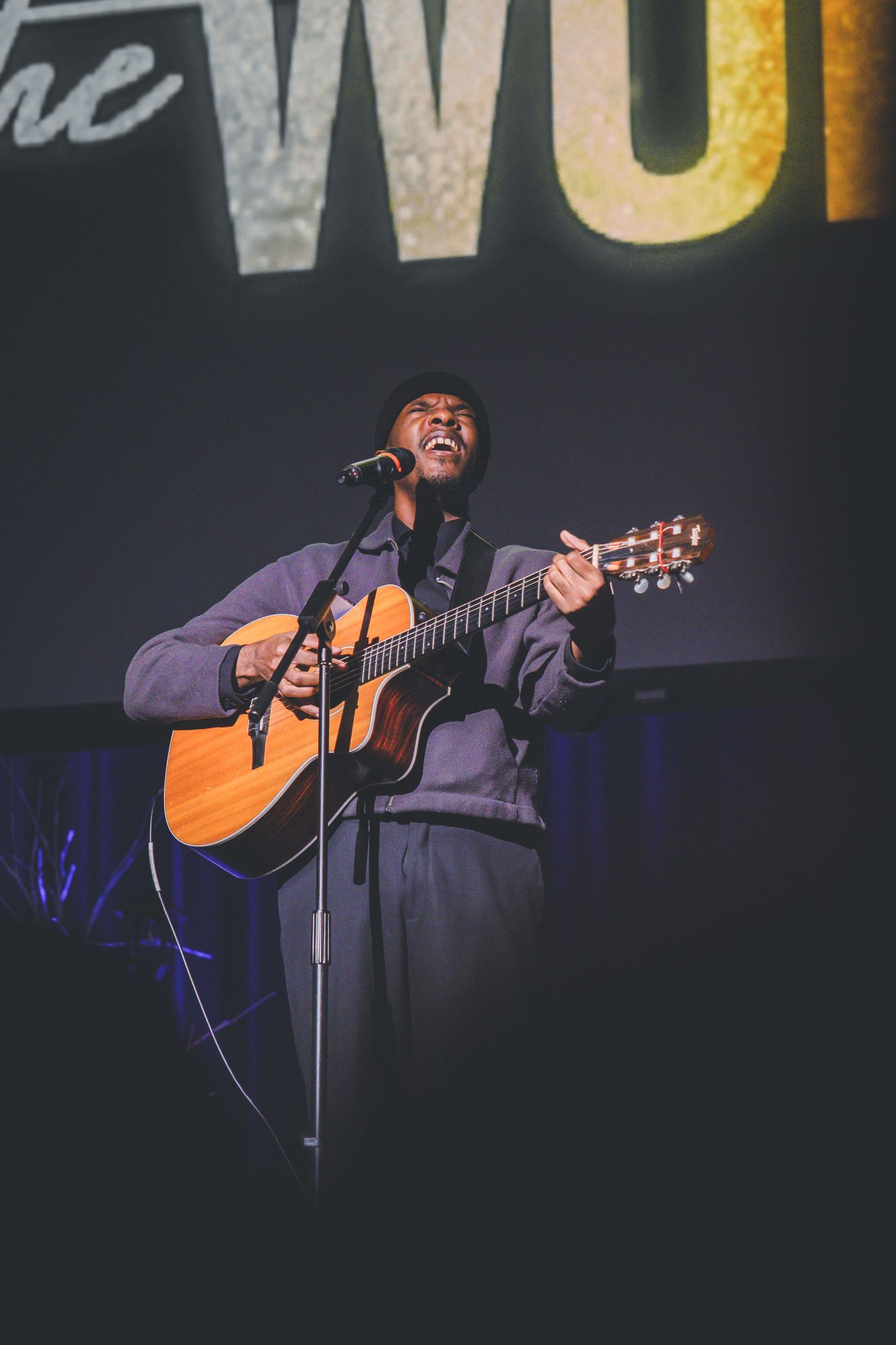 Musician plays acoustic guitar while performing on stage with illuminated text backdrop.