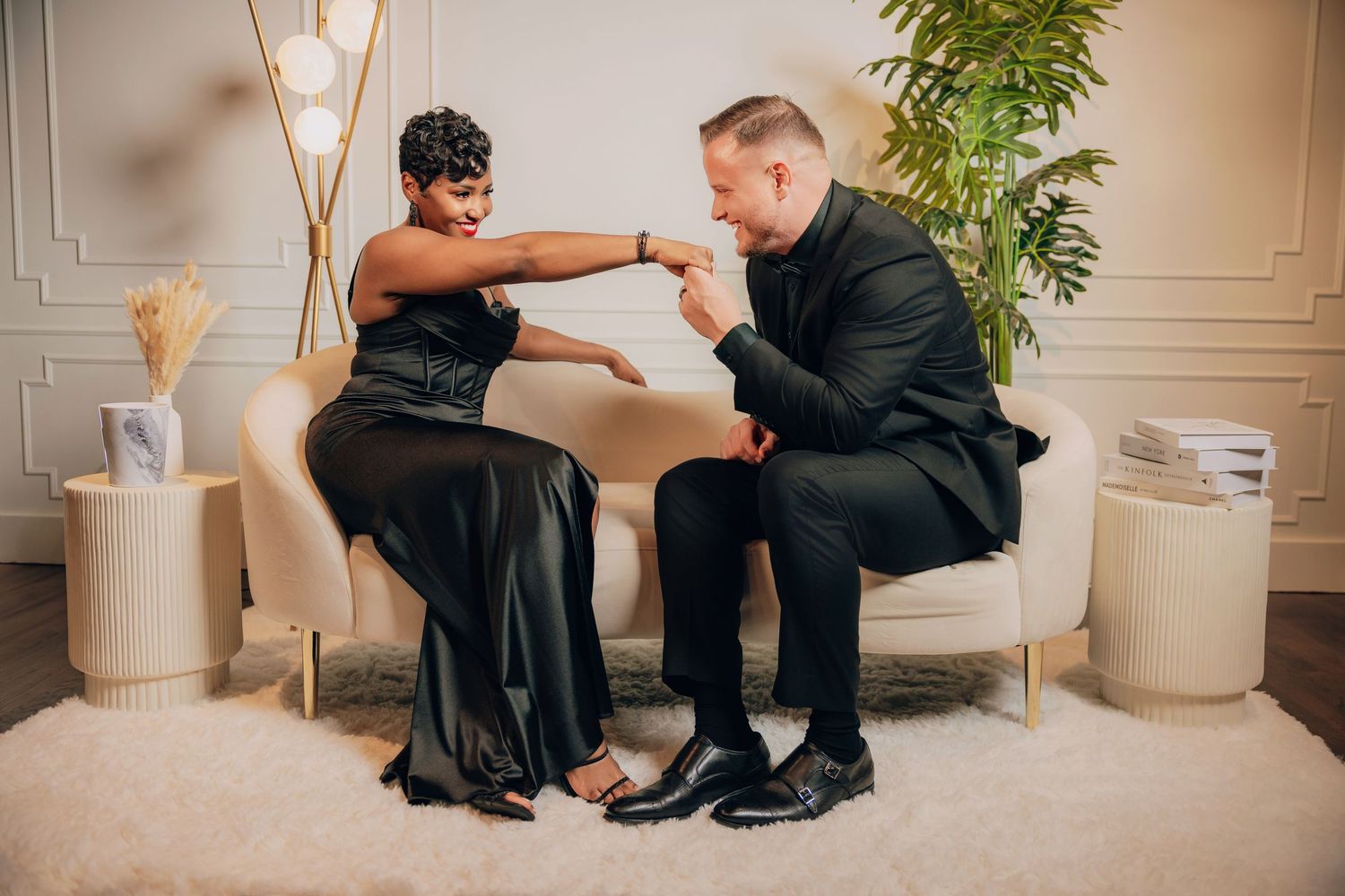 A person in a black evening gown sits on a white sofa with another person in a dark suit in an elegant setting.