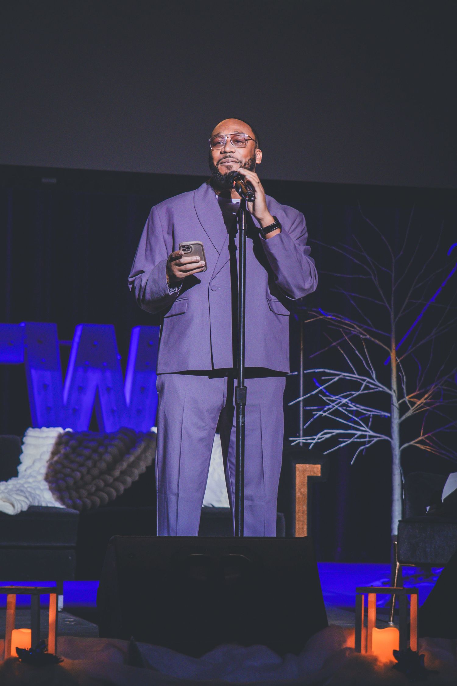 Performer in purple outfit stands on stage with blue lighting and decorative branches.