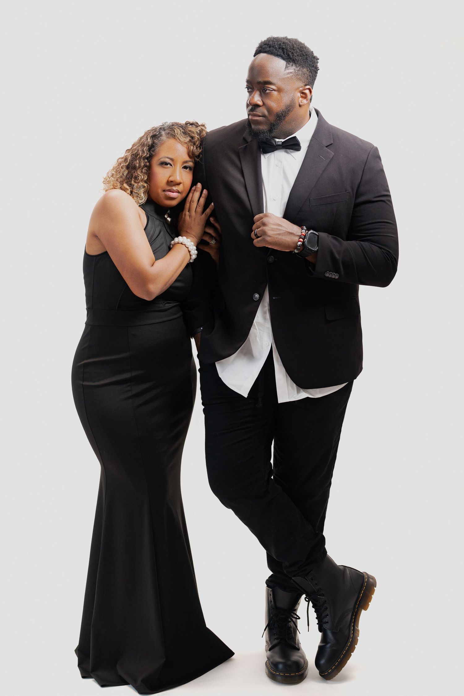 Couple in formal black attire poses intimately against white background in romantic photoshoot series.