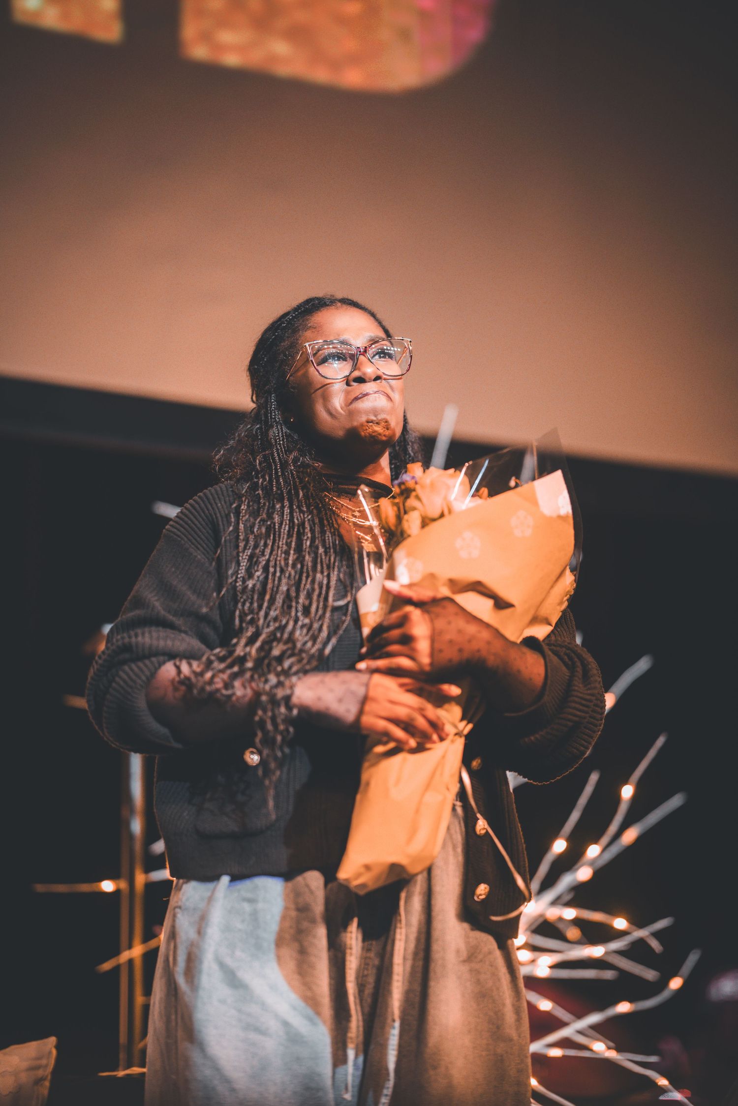 Person with dreadlocks and glasses holds an orange bouquet during a performance.