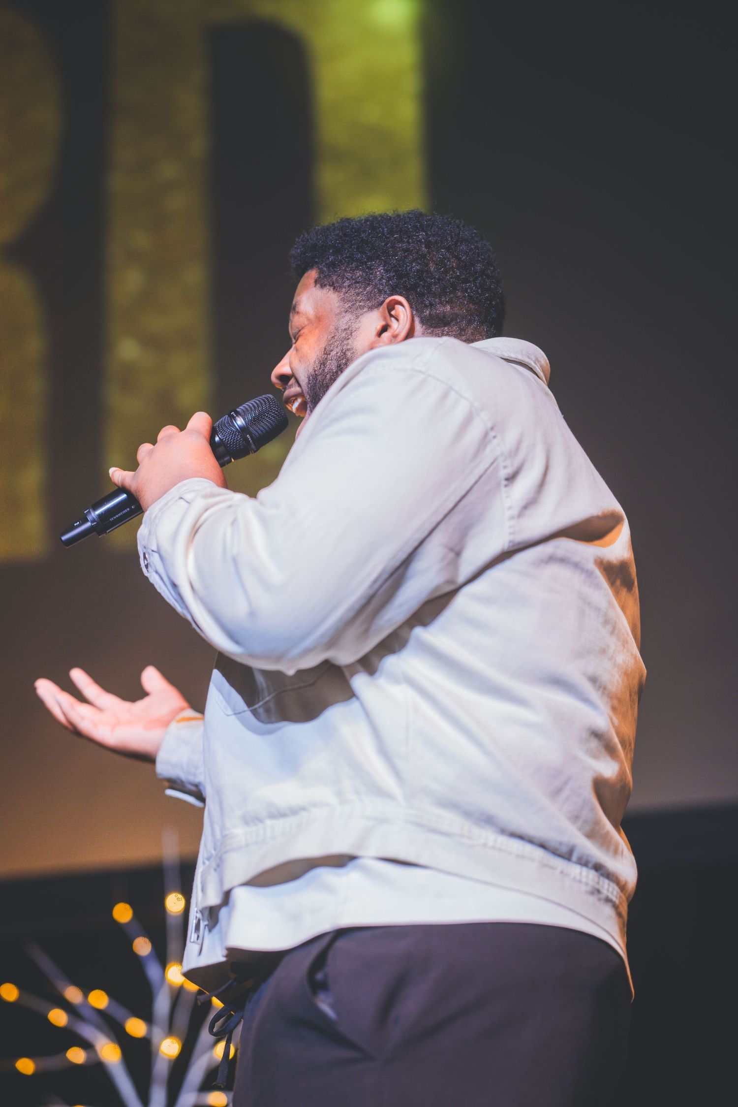 Performer in light gray jacket gestures while singing into microphone on stage.
