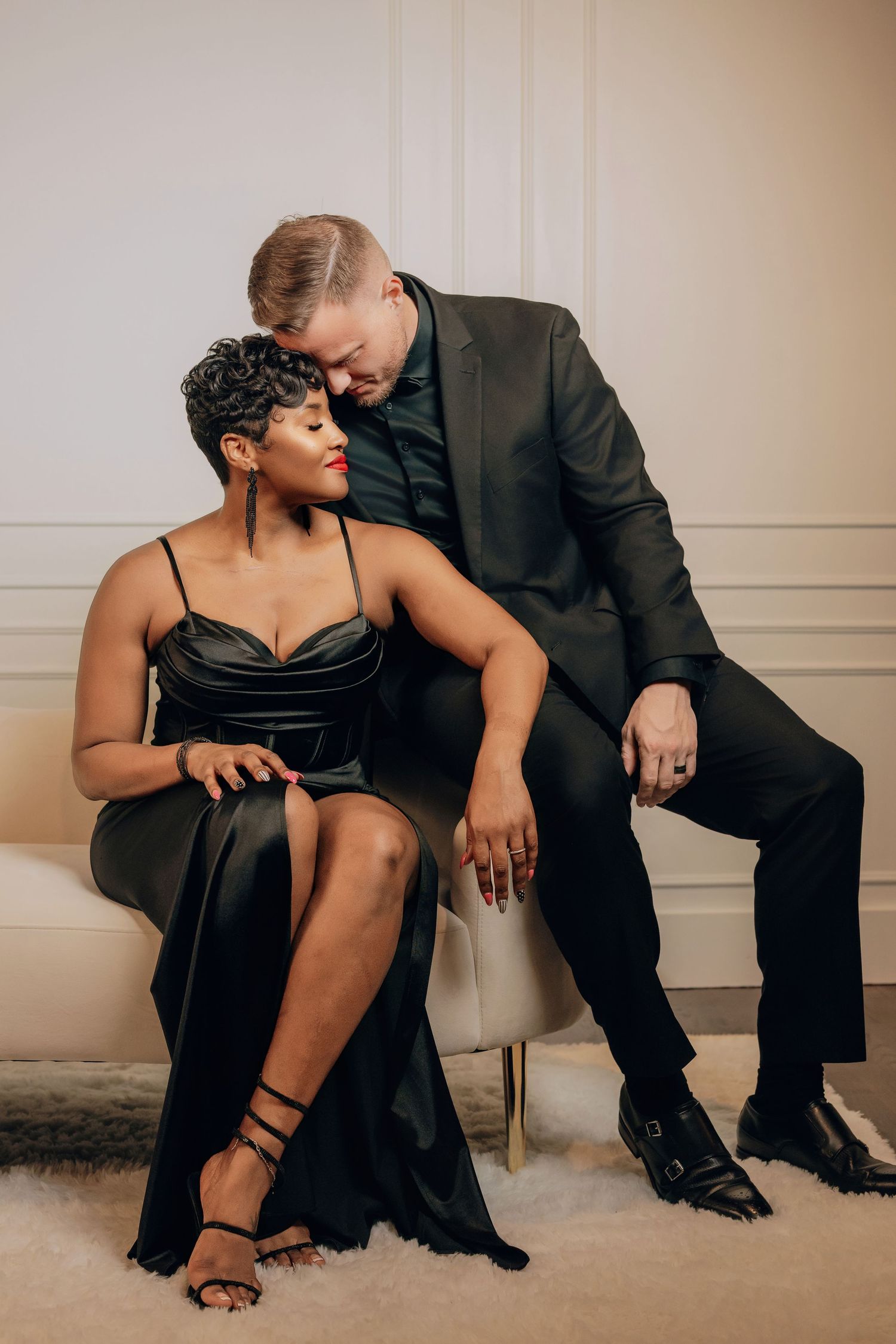 A romantic couple in formal black outfits share an intimate moment while seated on a light-colored chair.