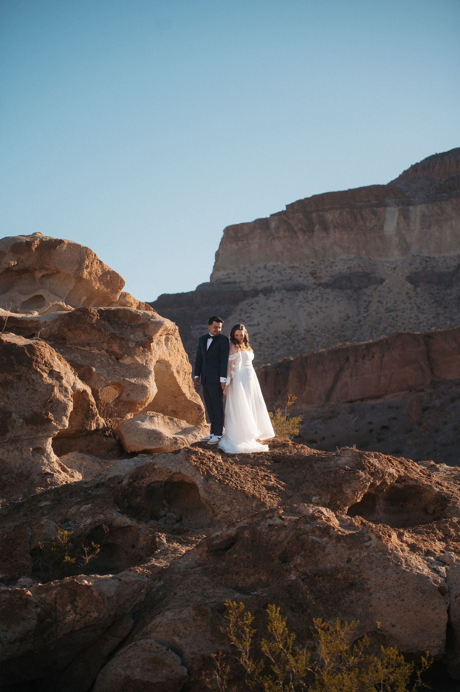 Bride and groom stand together on rugged desert rocks with dramatic canyon cliffs and blue sky behind them. Corpus Christi Wedding