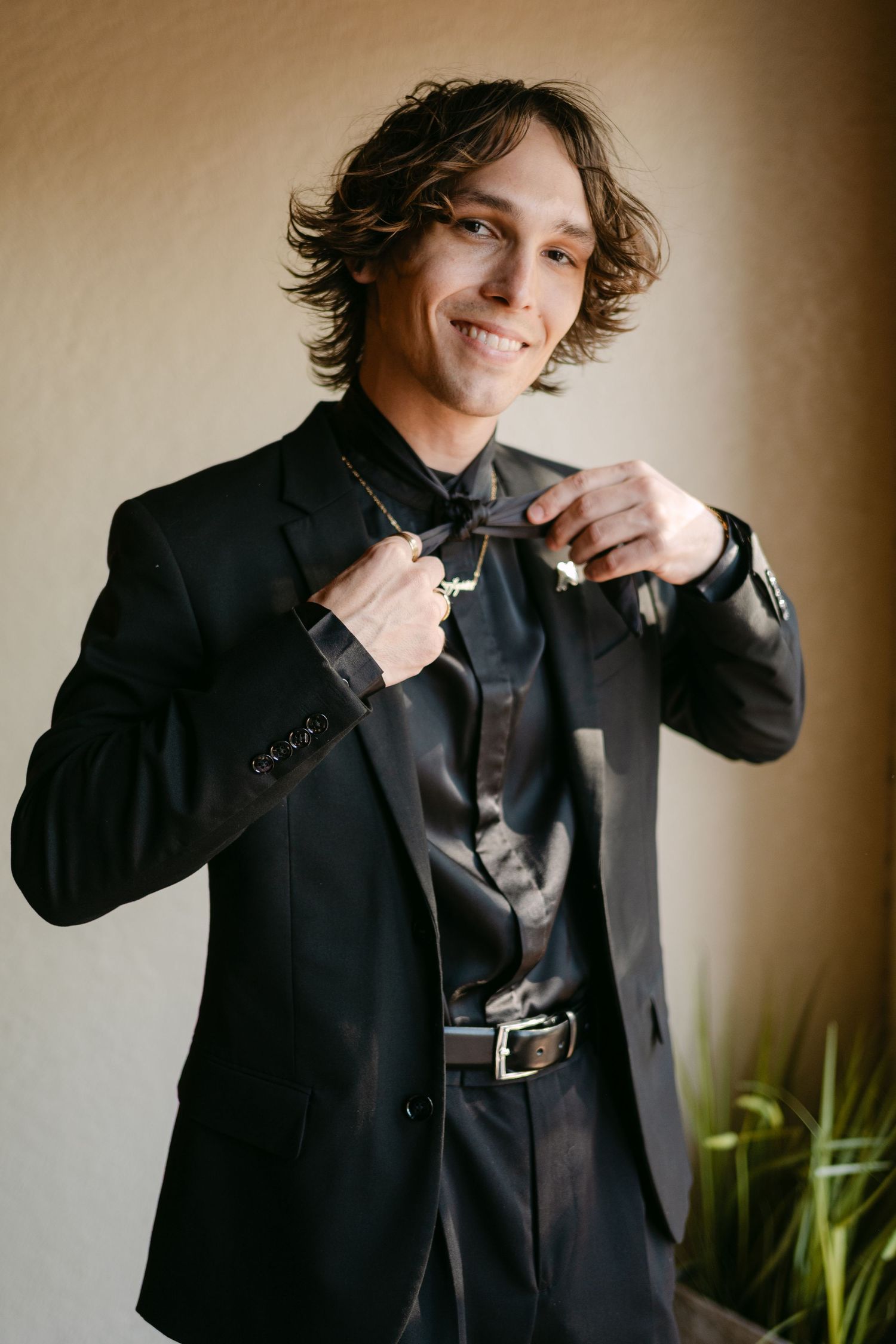 Smiling groom with curly hair in all-black suit adjusting bolo tie before wedding ceremony. Corpus Christi Wedding Photographer