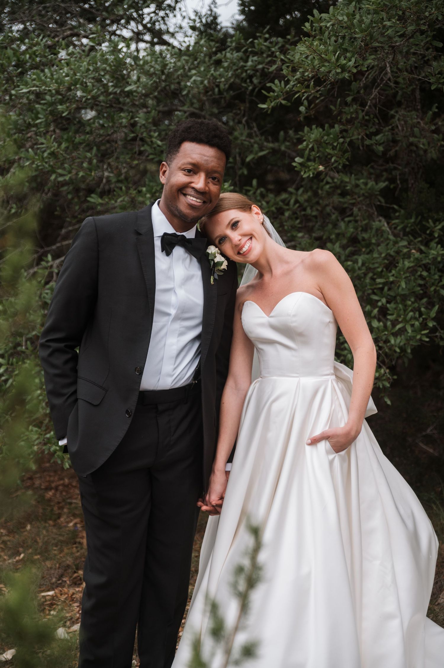 Smiling interracial couple poses together in wedding attire surrounded by lush green trees. Corpus Christi Wedding Photographer