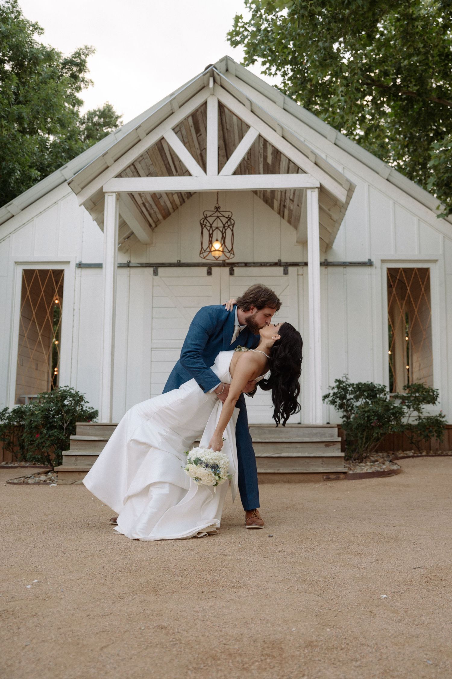 Groom dips bride for a kiss in front of a charming white chapel surrounded by lush green trees. Corpus Christi Wedding Photographer