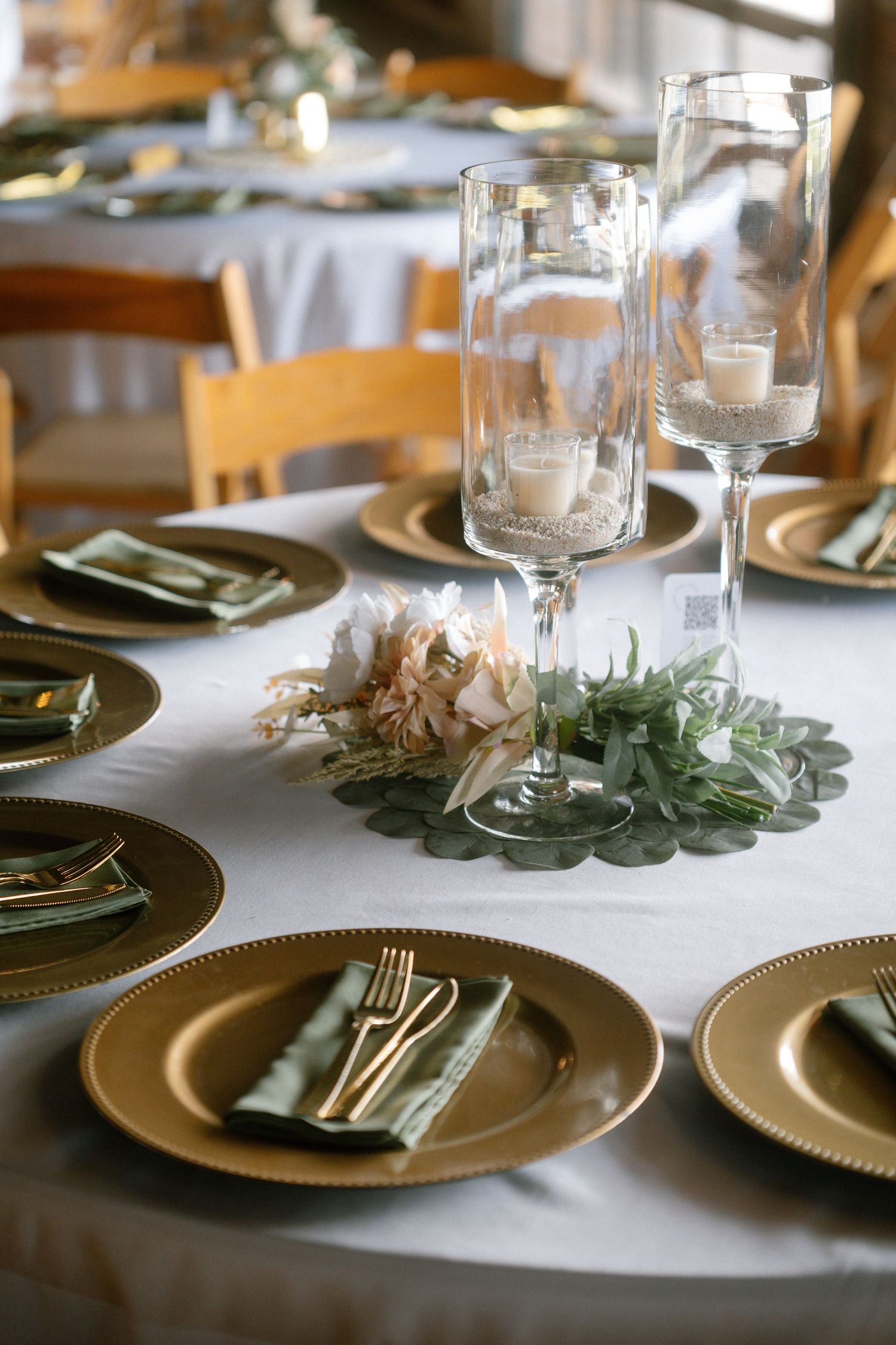 Elegant wedding reception table set with gold charger plates, green napkins, and glass candle centerpieces on white linen. Corpus Christi
