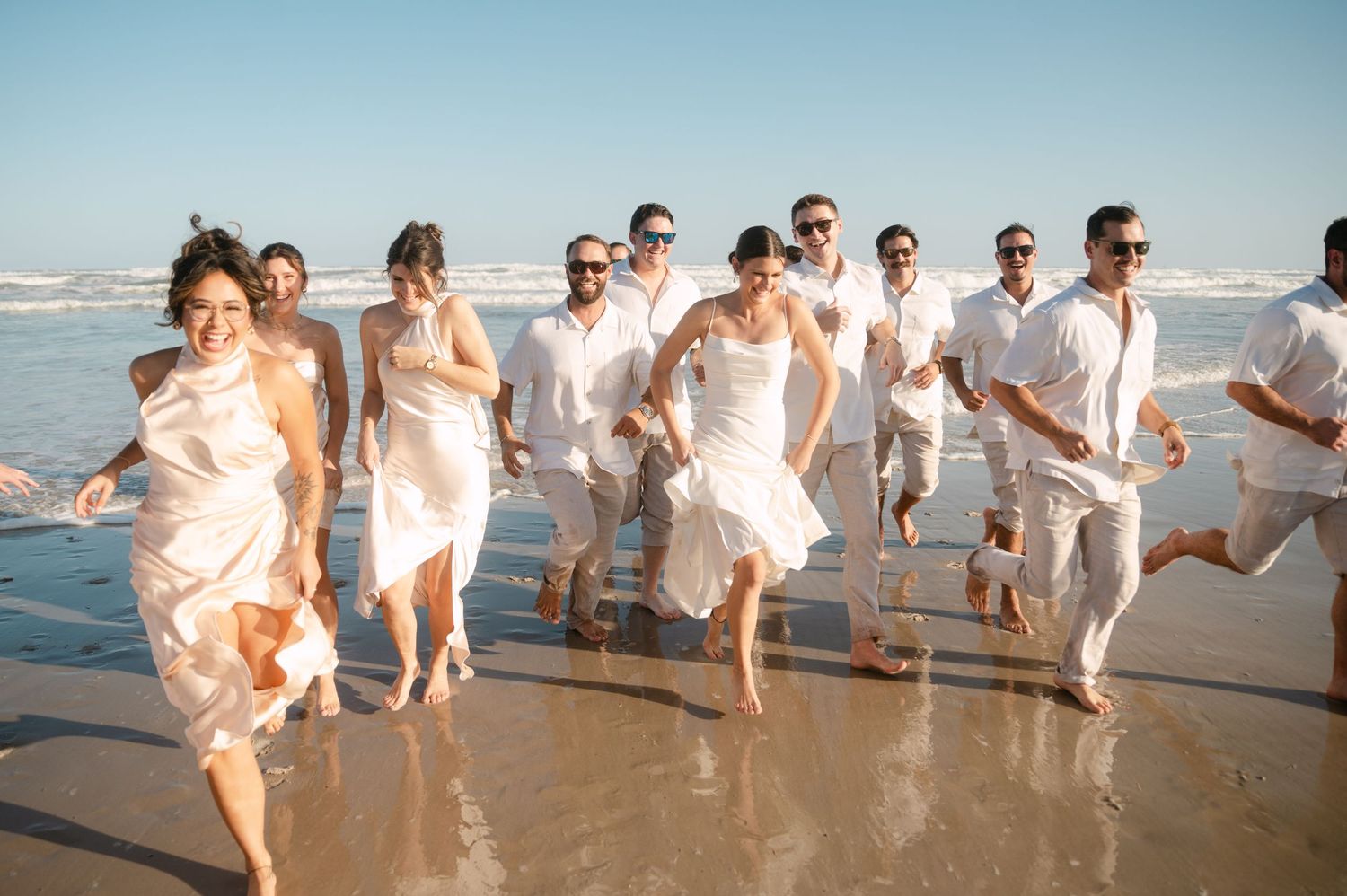 Wedding party in matching white outfits running joyfully along sunny ocean beach shoreline. Corpus Christi Wedding Photographer