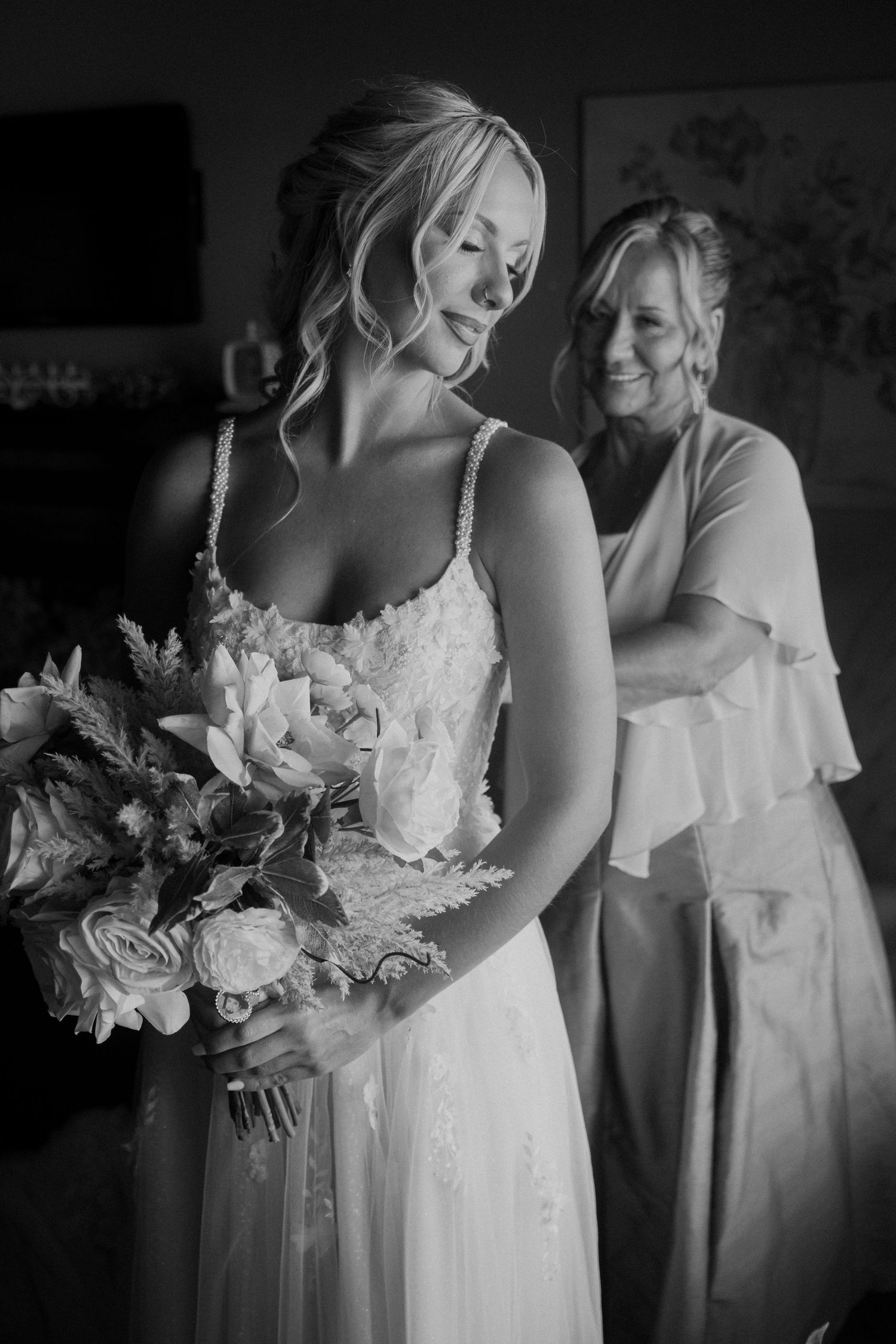 Black and white photo of smiling bride in lace gown holding bouquet while woman helps with dress behind her. Corpus Christi Wedding Photogra
