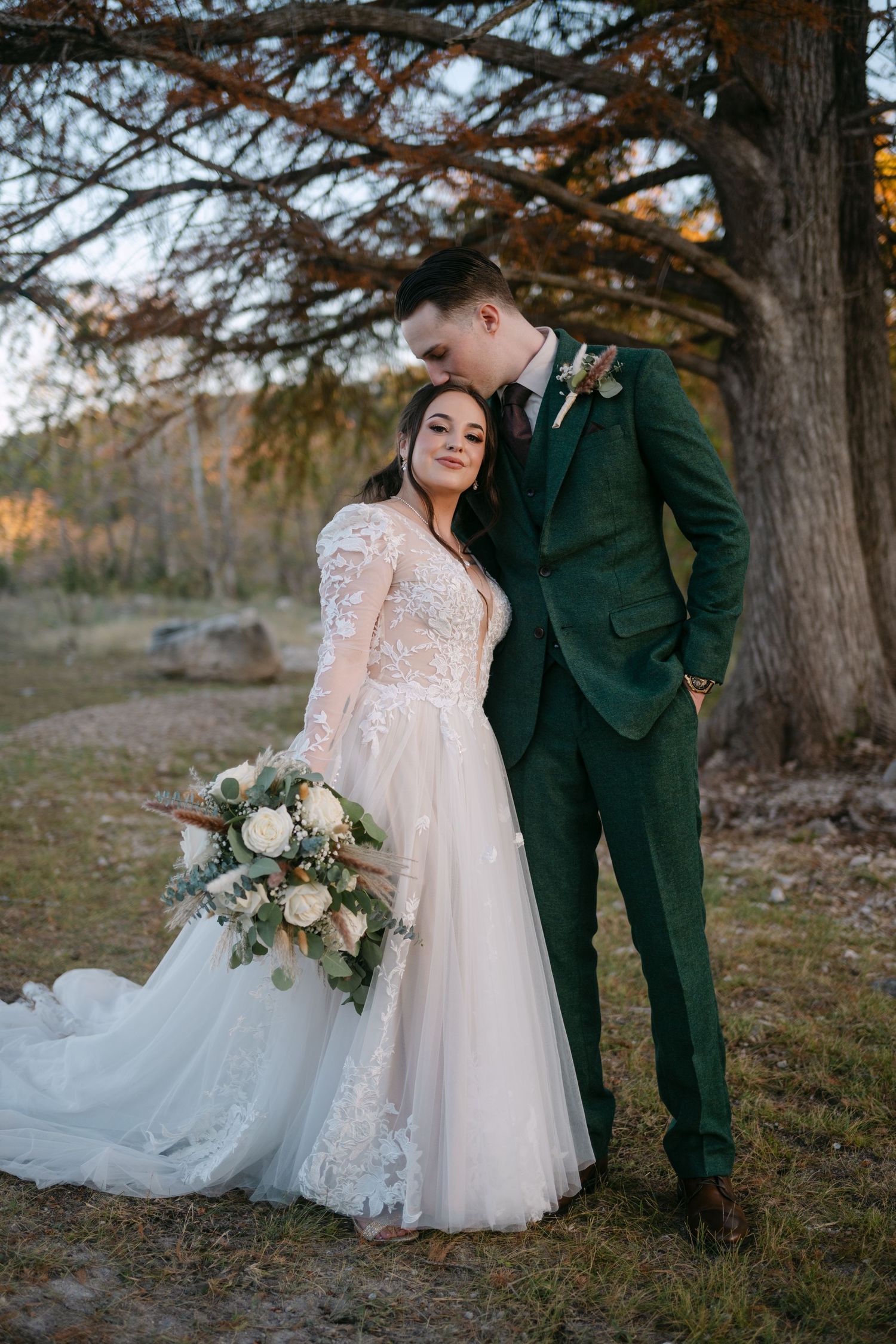 Bride in lace gown and groom in green suit embrace tenderly under a large autumn tree at golden hour. Corpus Christi Wedding Photographer