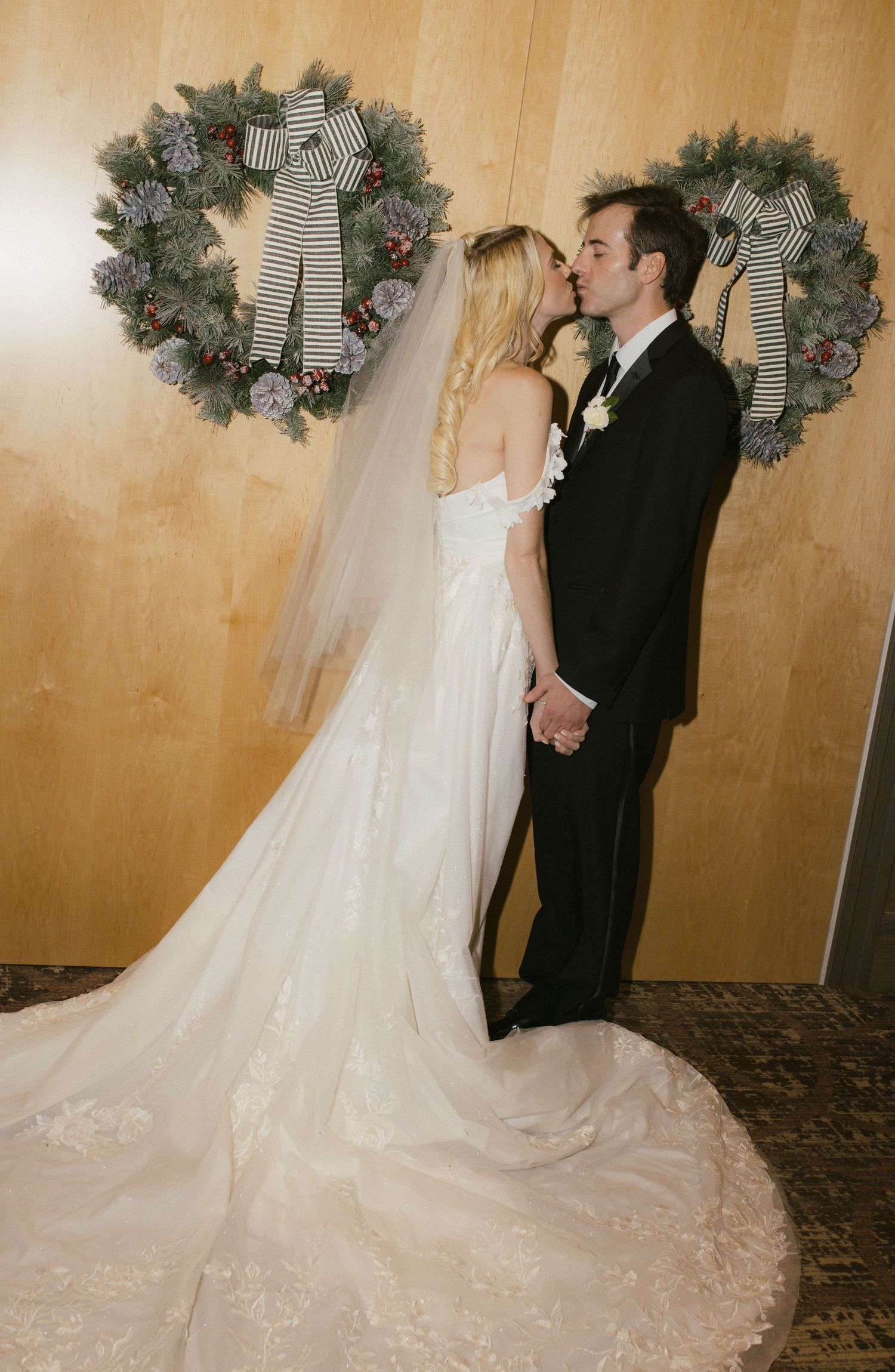 A wedding couple shares a kiss in front of holiday wreaths with striped bows.
