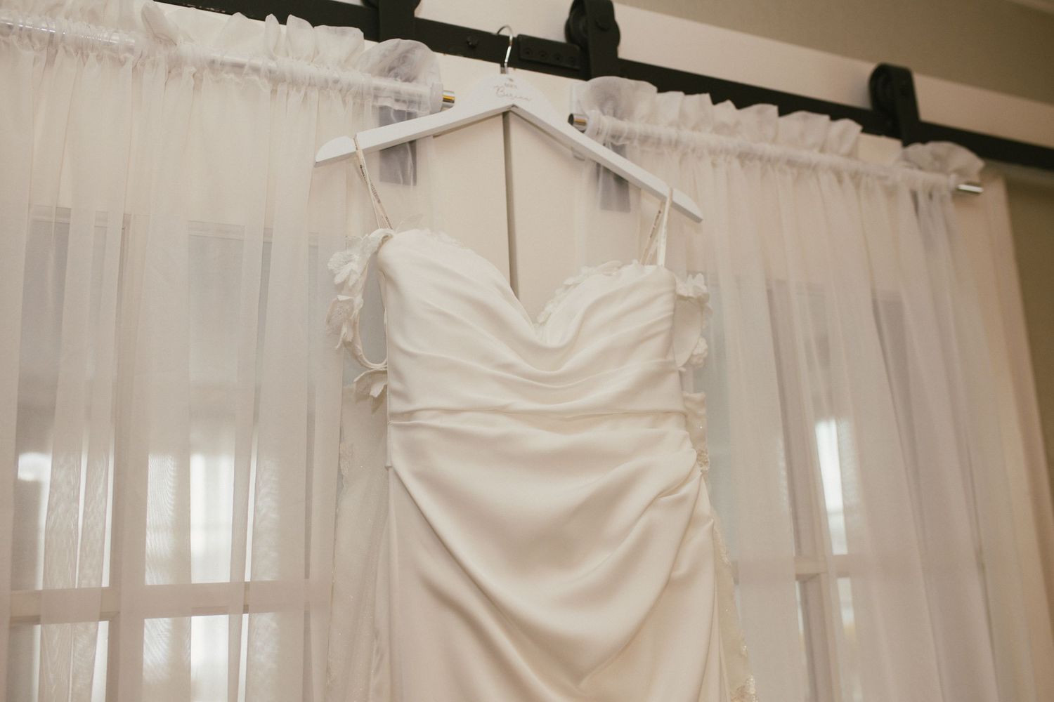 Wedding dress displayed on a hanger against white curtains in natural light.