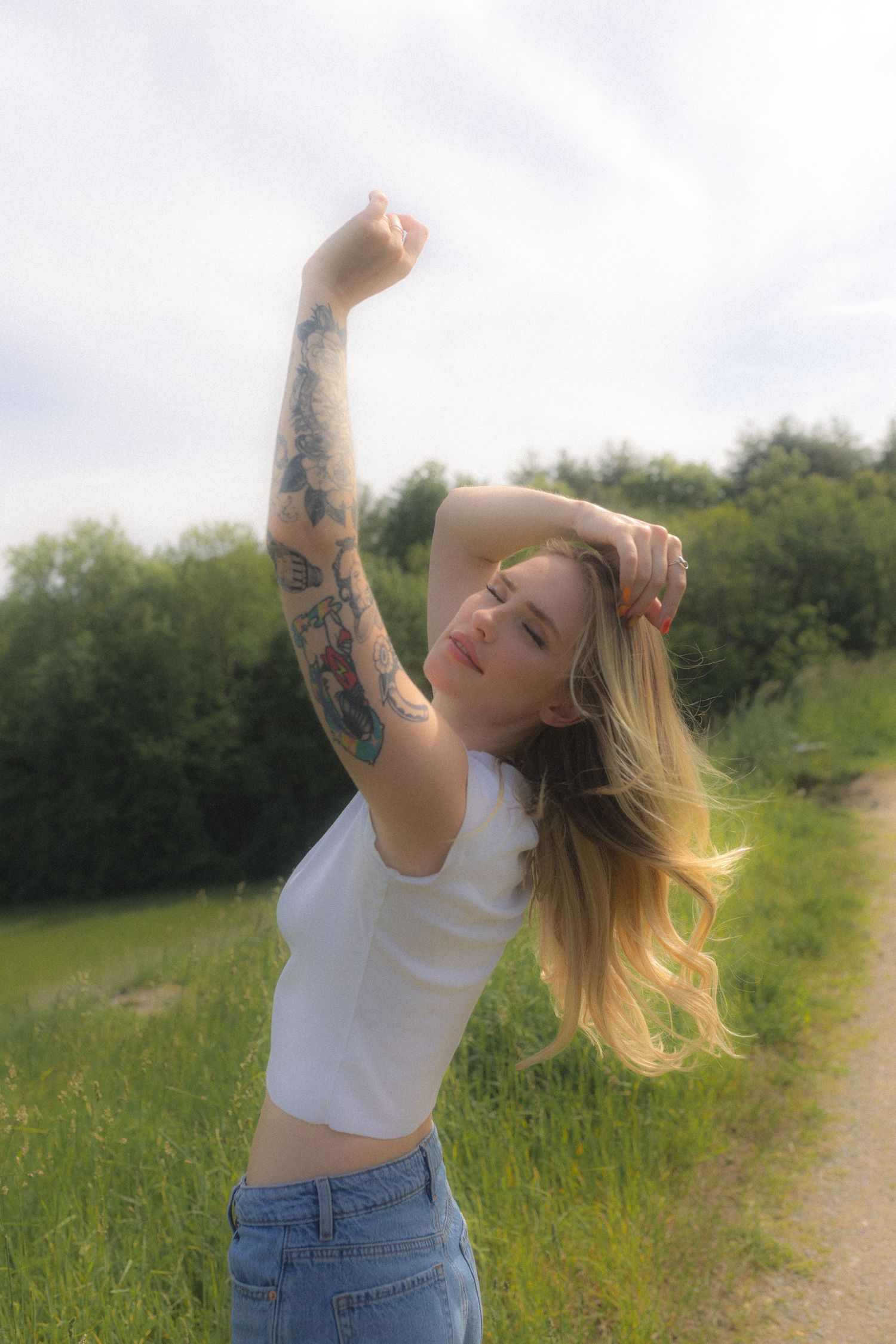 A person with tattoos dances freely on a sunny path surrounded by tall grass.
