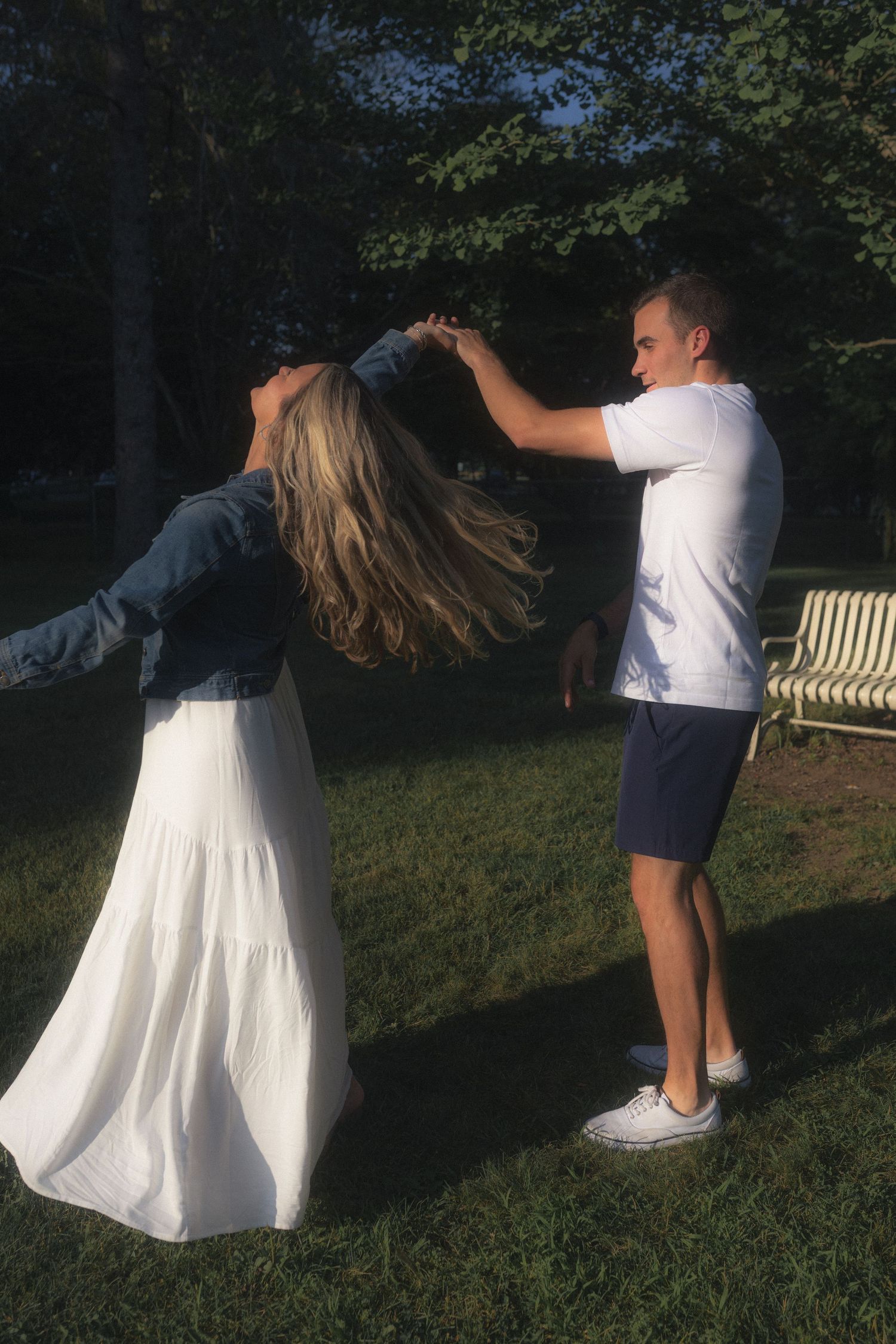 A couple in white and blue outfits dancing together on grass in a park setting.