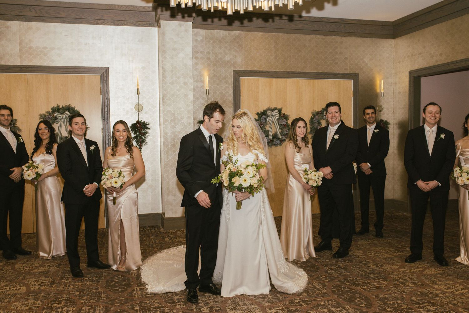 Wedding party poses together in formal attire with bridesmaids in champagne dresses and groomsmen in black suits.