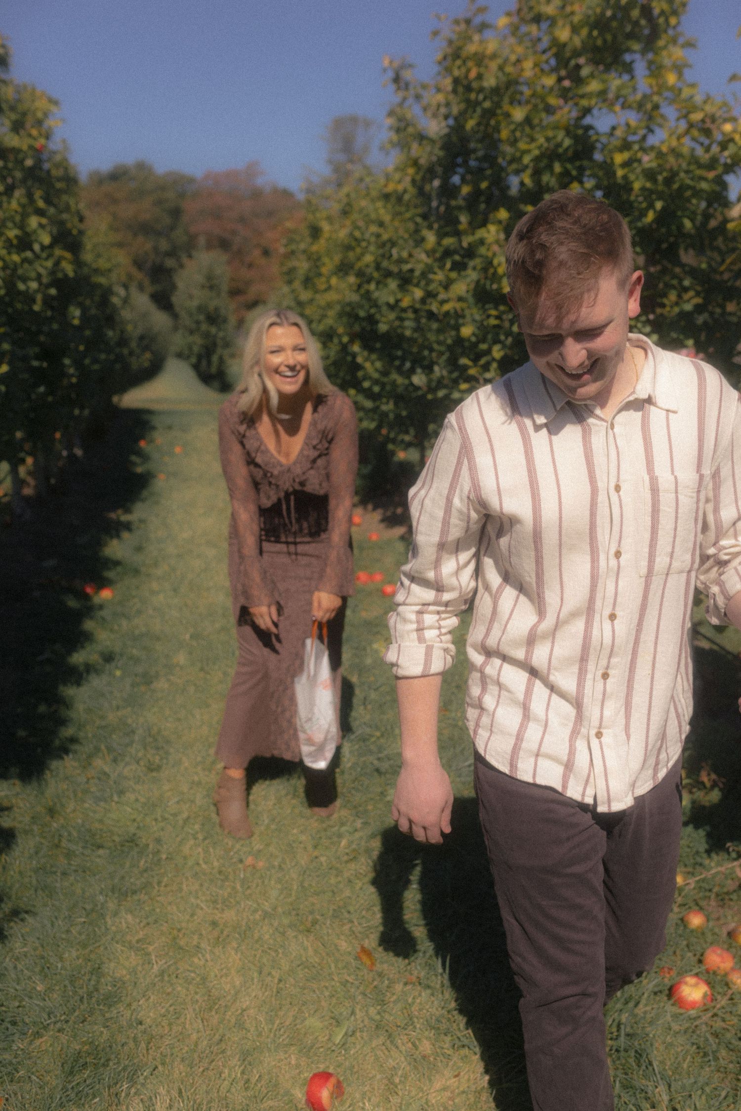 A playful couple walks through rows of apple trees in an orchard.