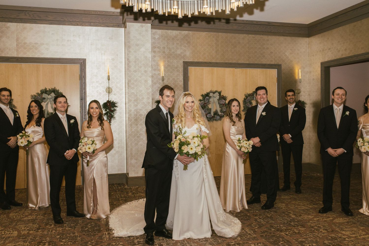 Formal wedding party portrait in an elegant venue with chandelier and neutral decor.