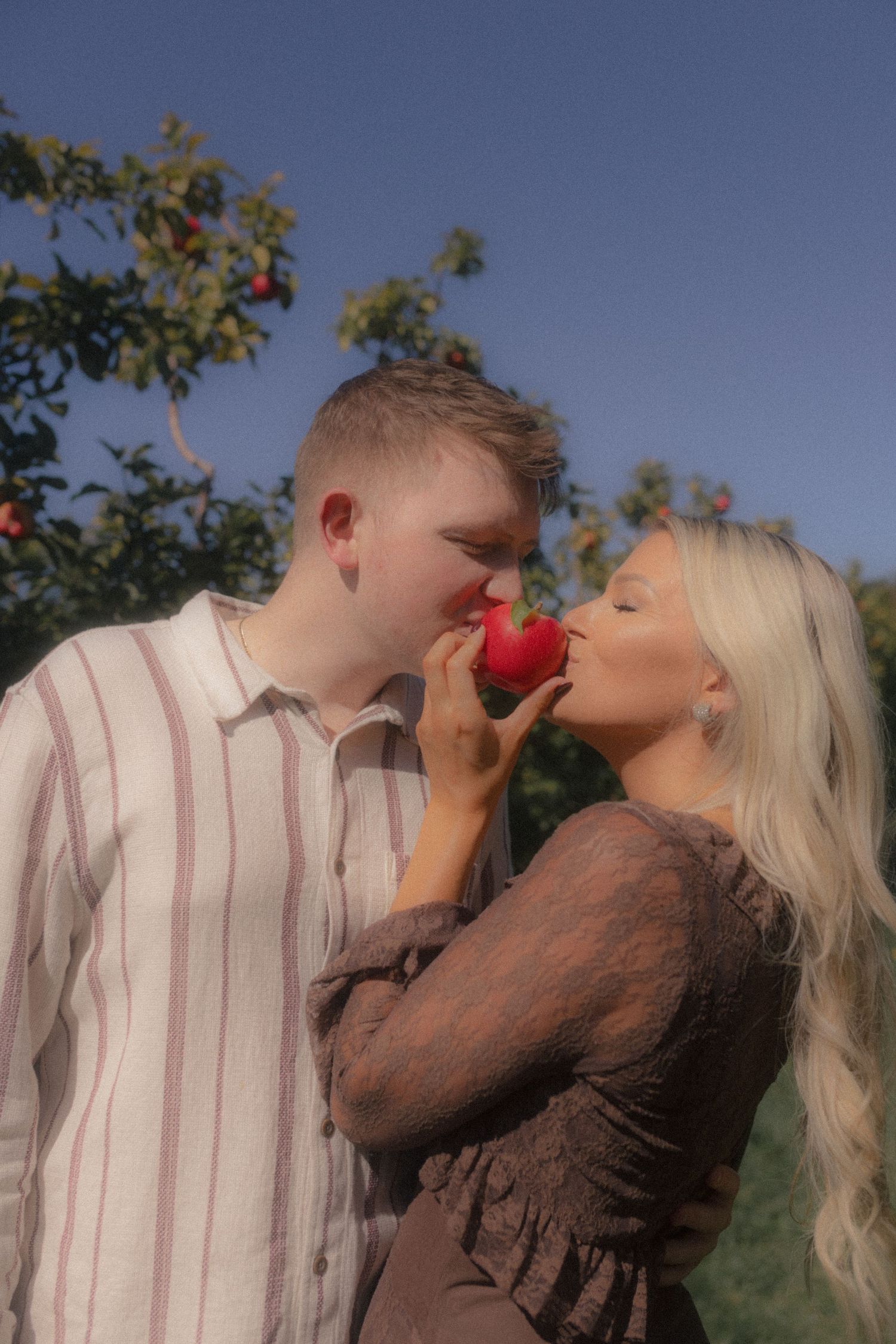 A couple poses playfully in an apple orchard on a sunny day.