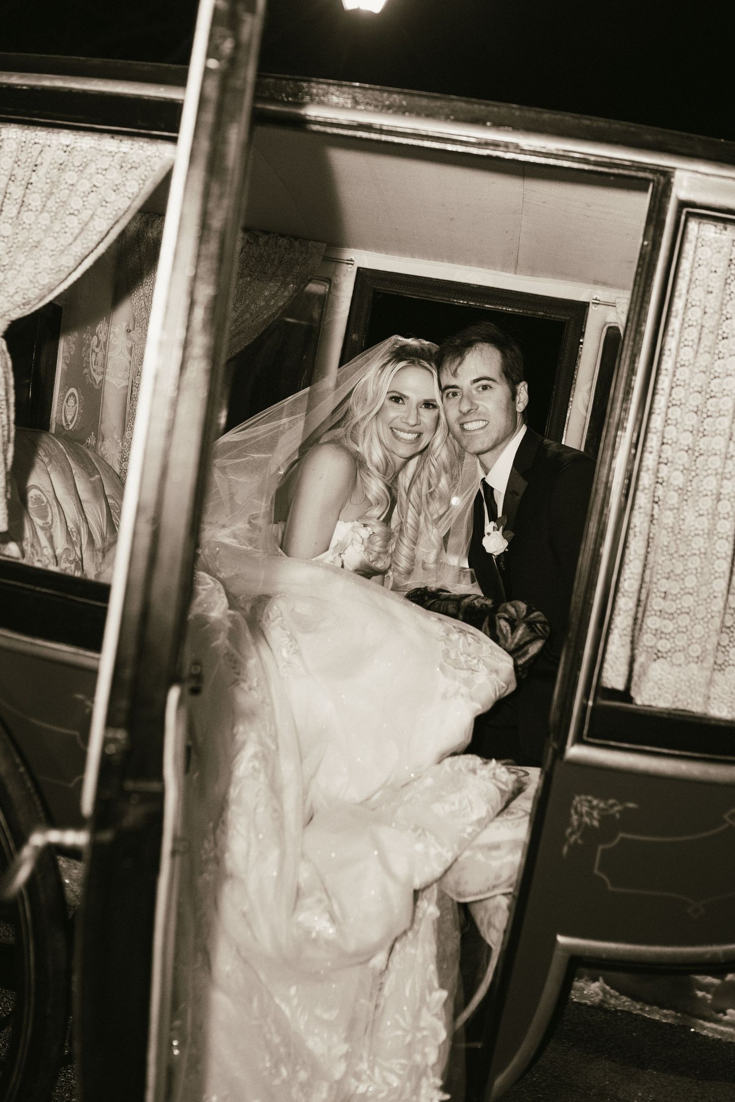 Newlyweds exit their wedding reception through a vintage car door in a black and white photograph.