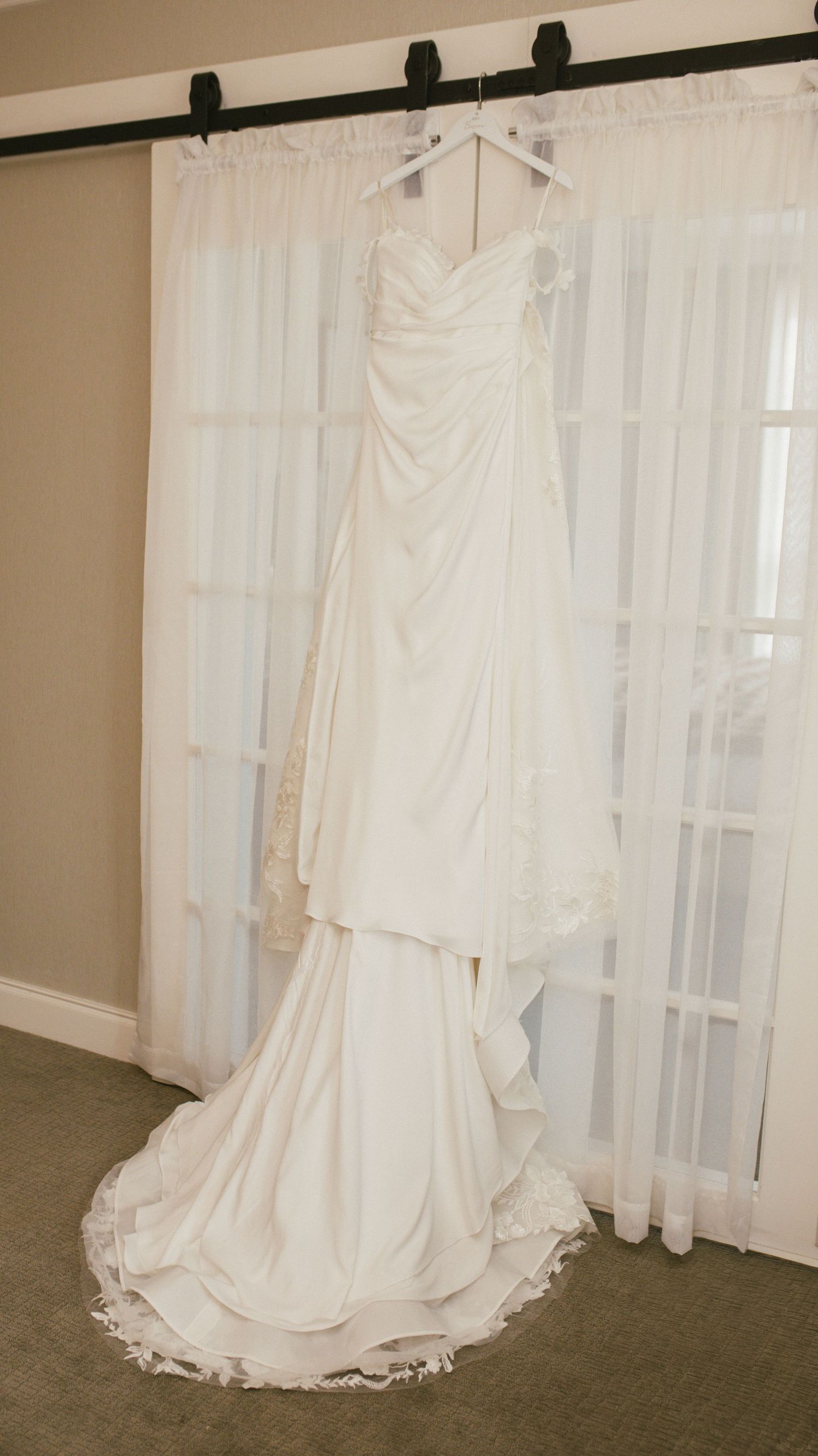 White wedding dress with flowing train displayed against sheer curtains in natural light.