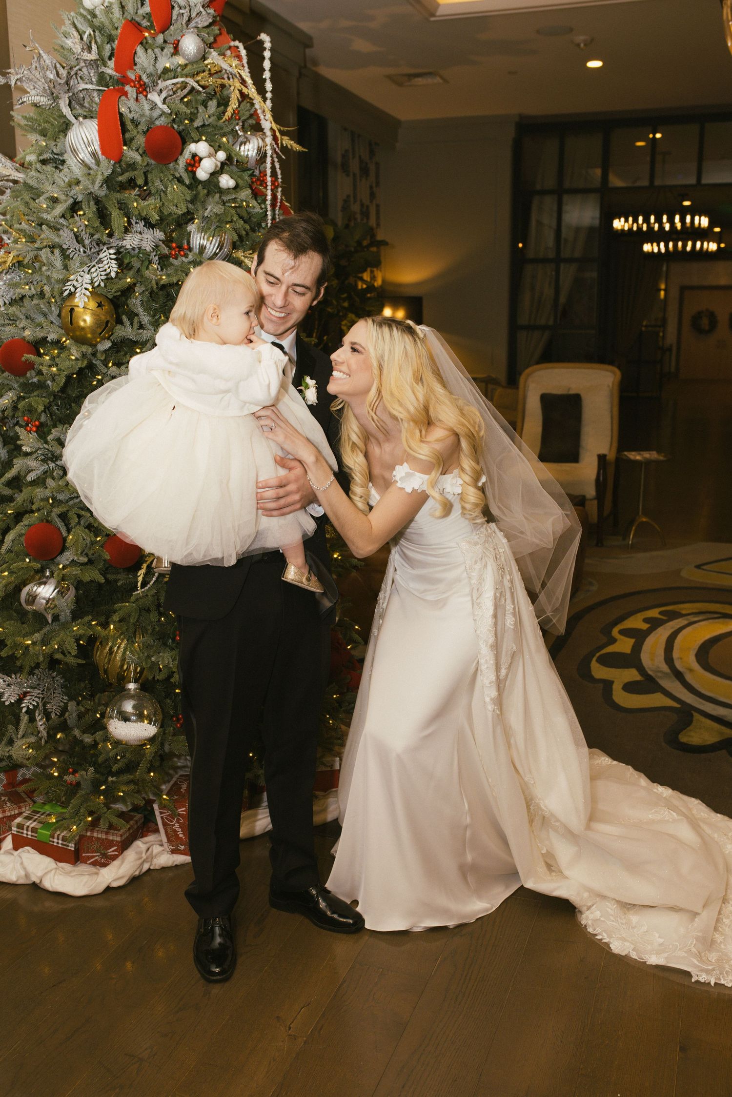 A wedding couple shares a moment with a small child by a decorated Christmas tree.