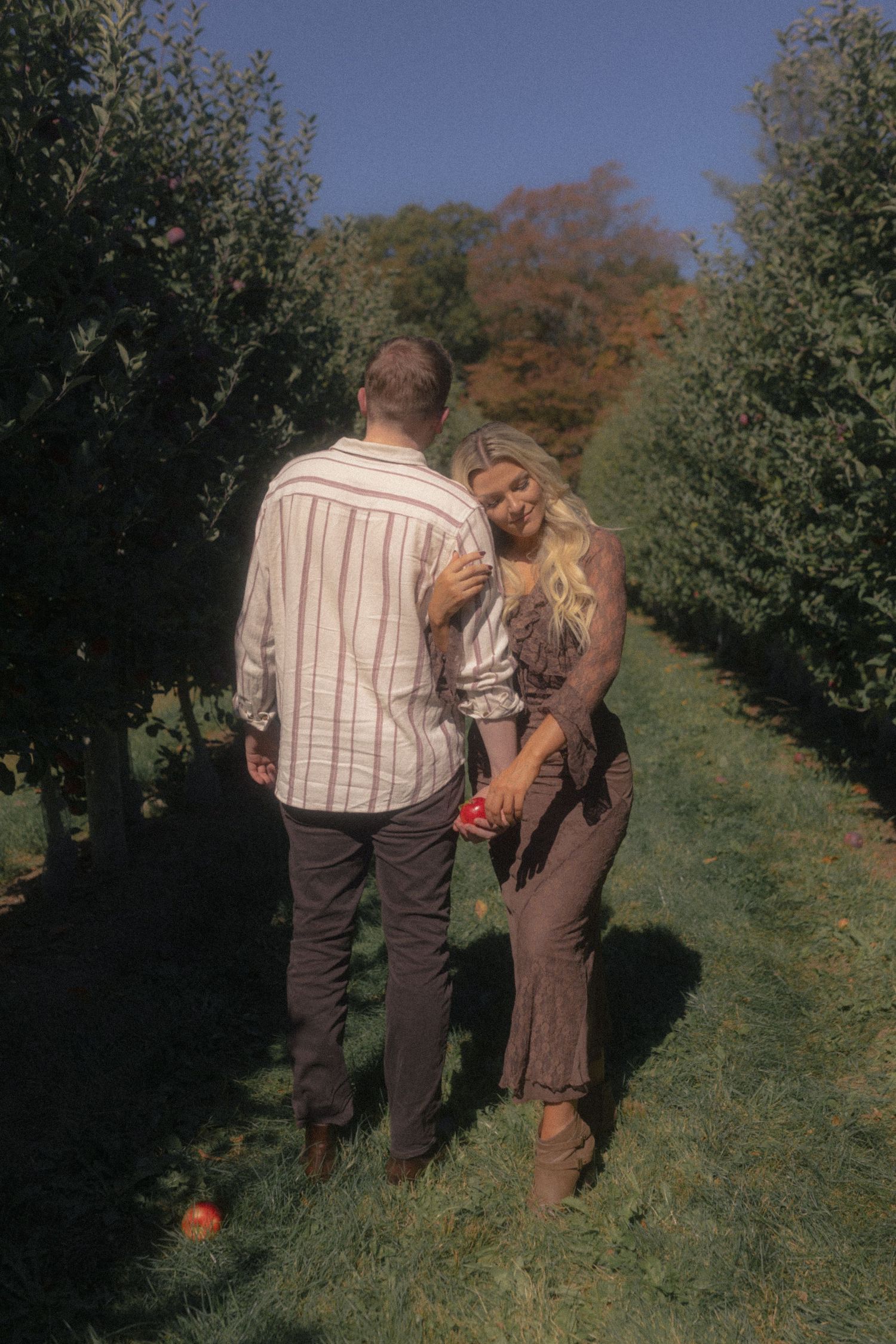 A couple strolls through an autumn orchard with pumpkins scattered along a grassy path.