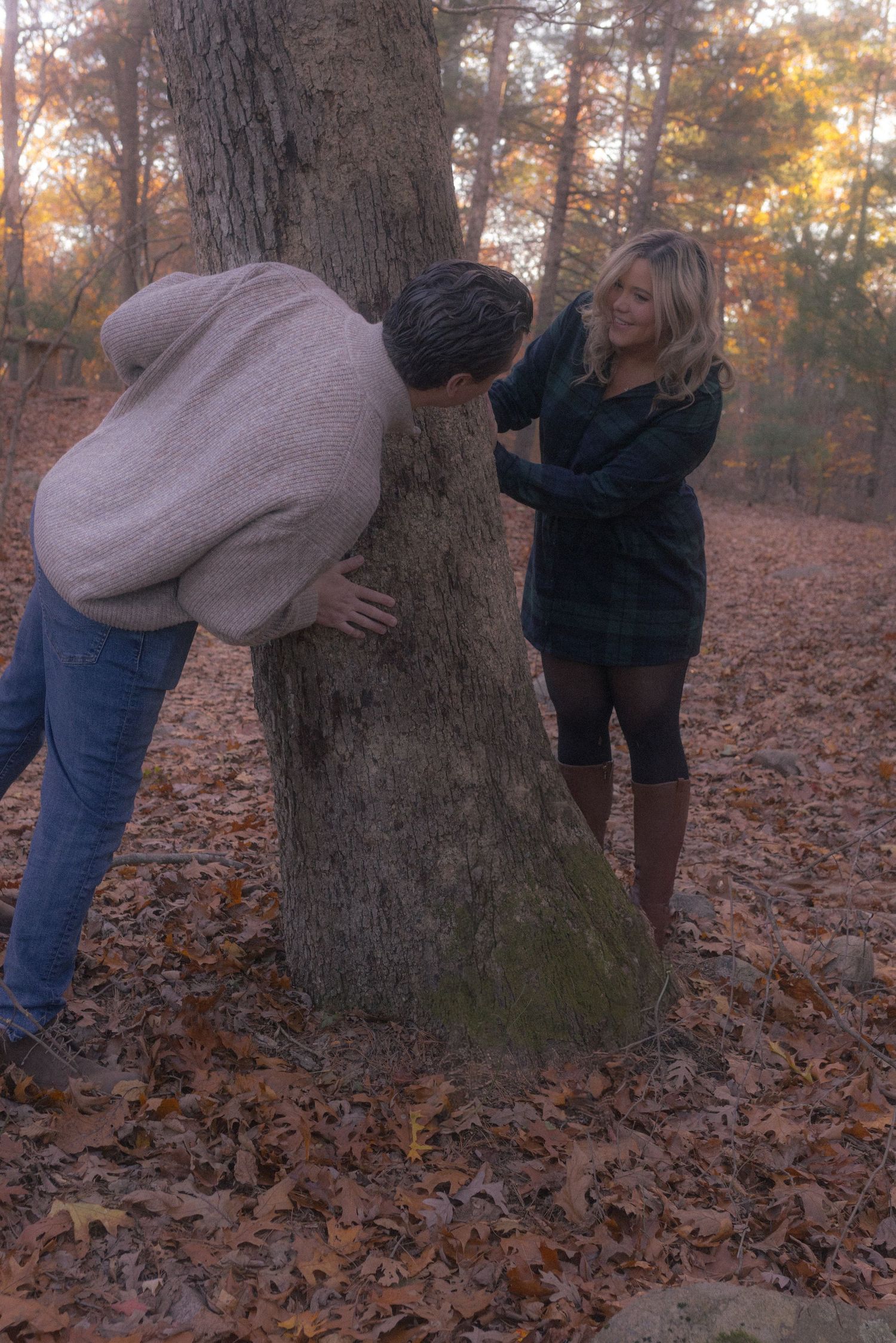 A playful autumn scene with a couple posing near a large tree trunk covered in fallen leaves.