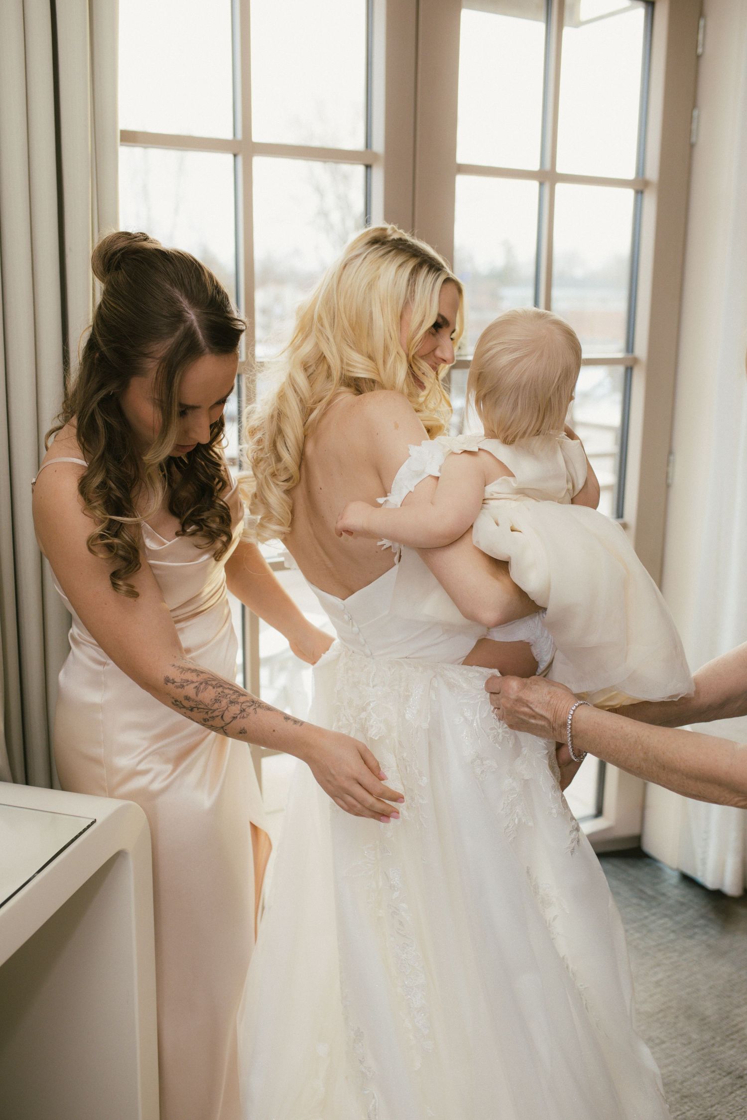 Bridesmaids in light pink dresses helping with a white wedding gown in a bright room.