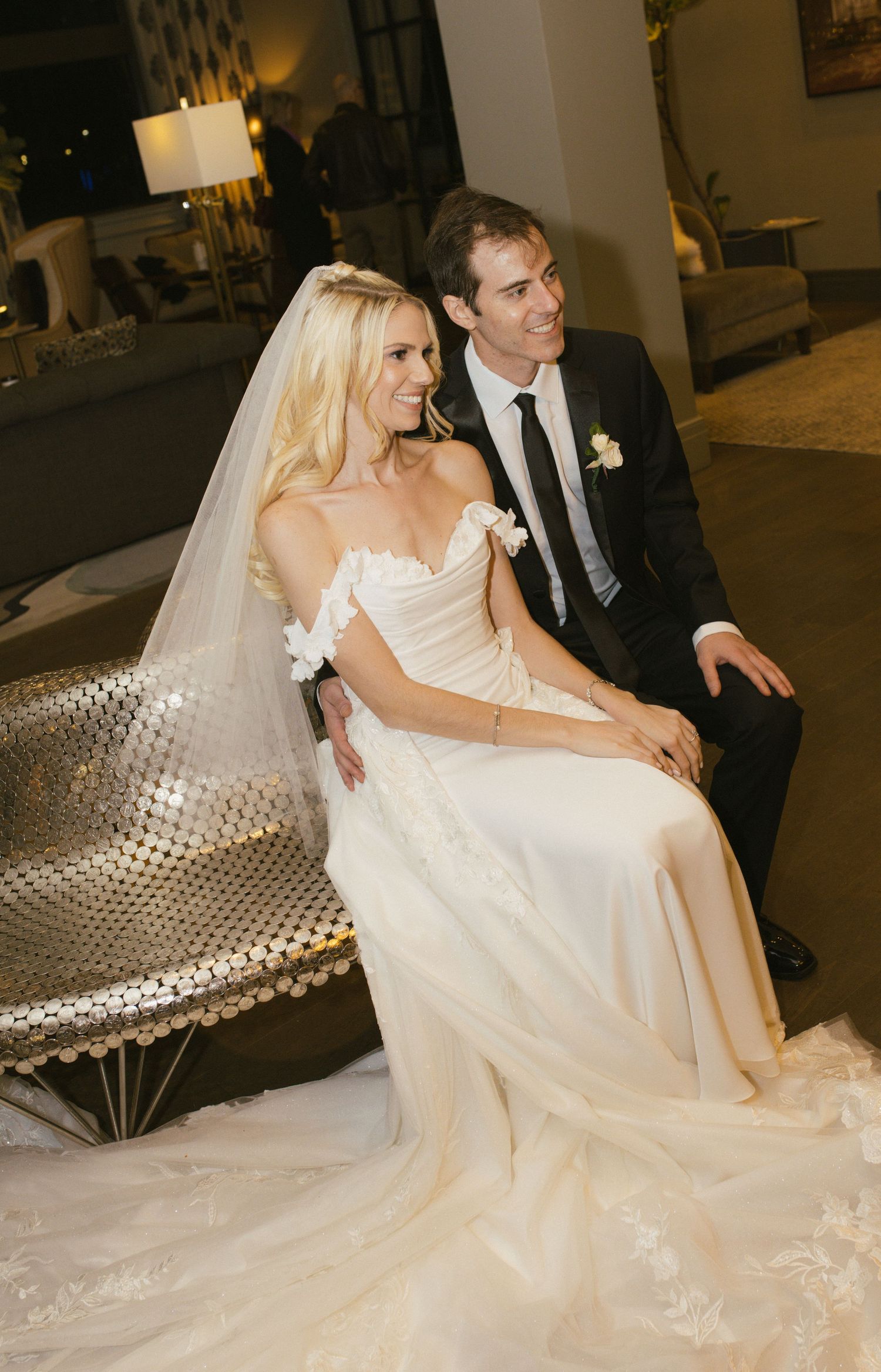 A bride in white gown sits with groom on metallic furniture at their reception.