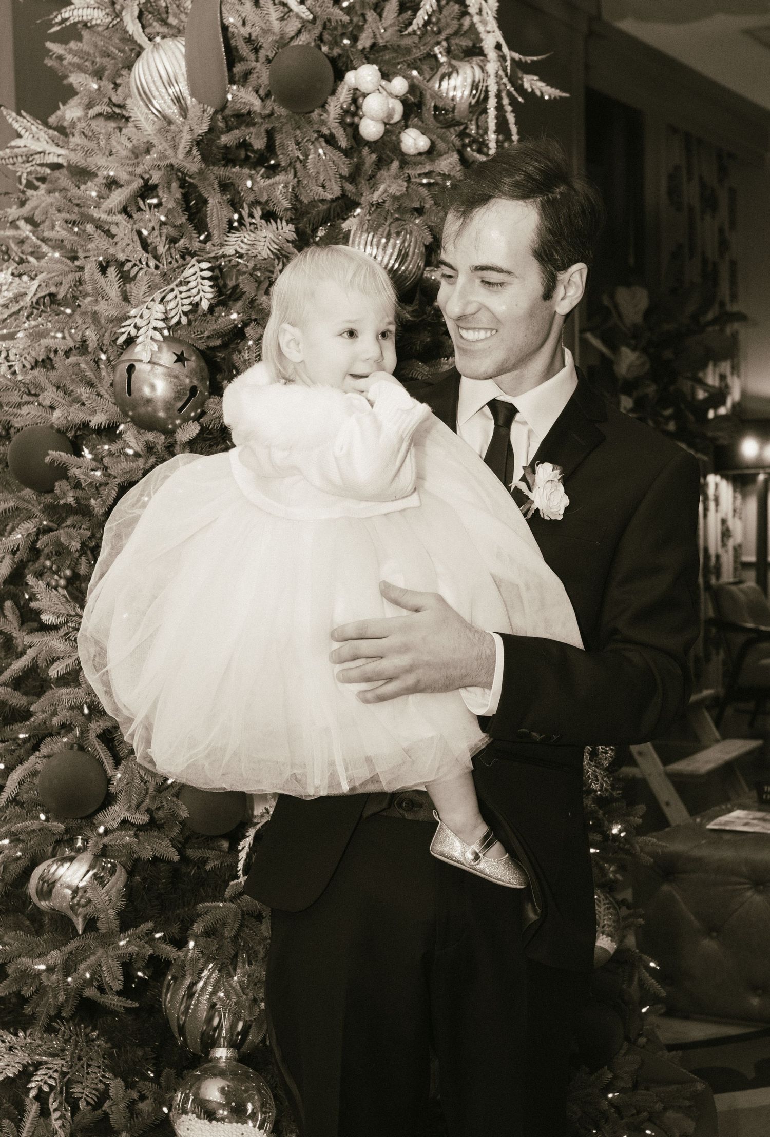 A formal portrait of a parent holding a baby in white dress by a Christmas tree.