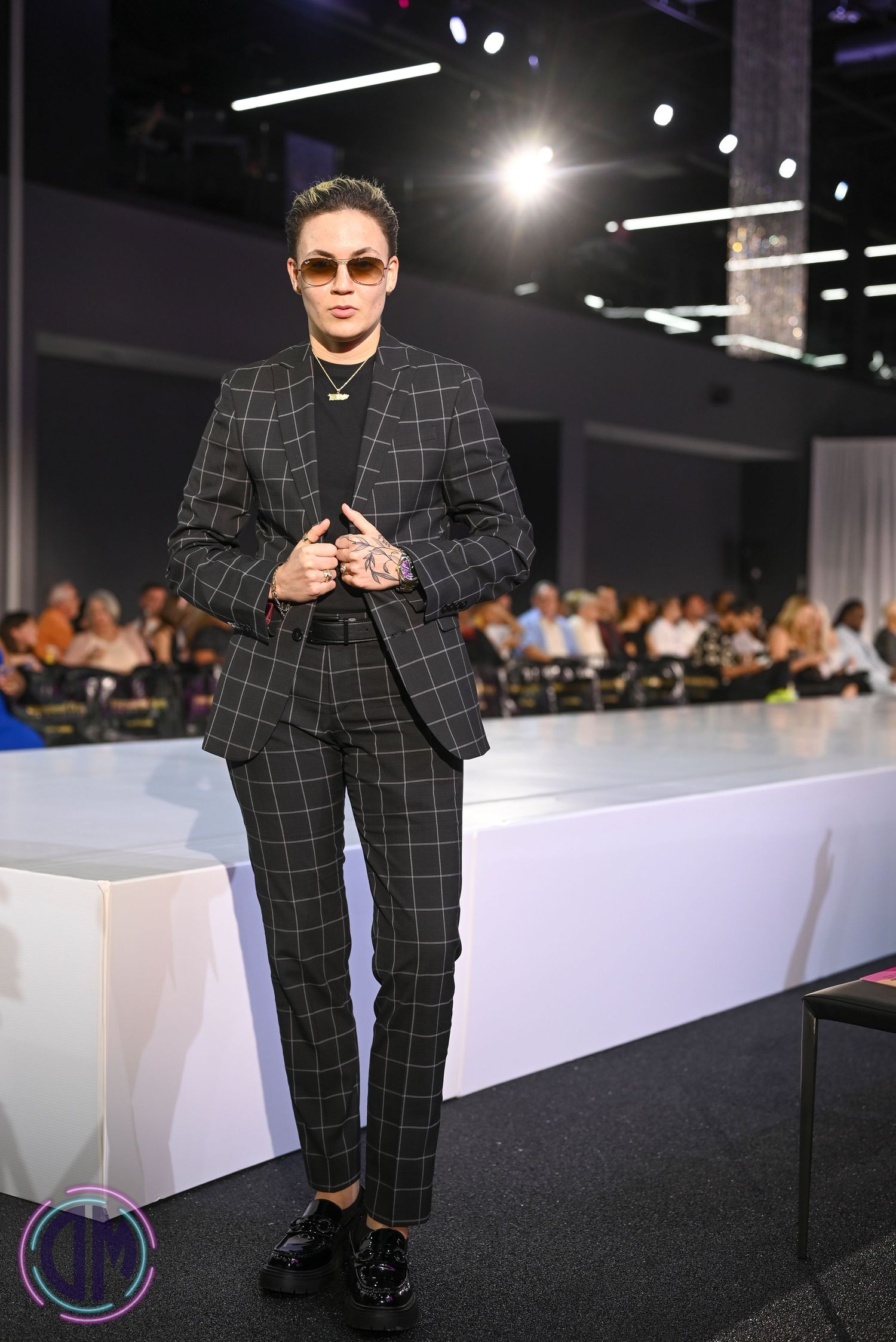 Checkered black suit ensemble being modeled on a dark runway with white barriers and audience seating.