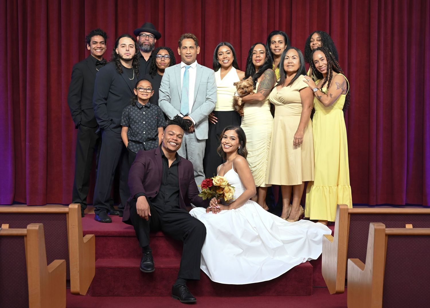 Bride in white gown holding a bouquet sits beside her groom as a group of family and friends pose on church steps before red curtains.