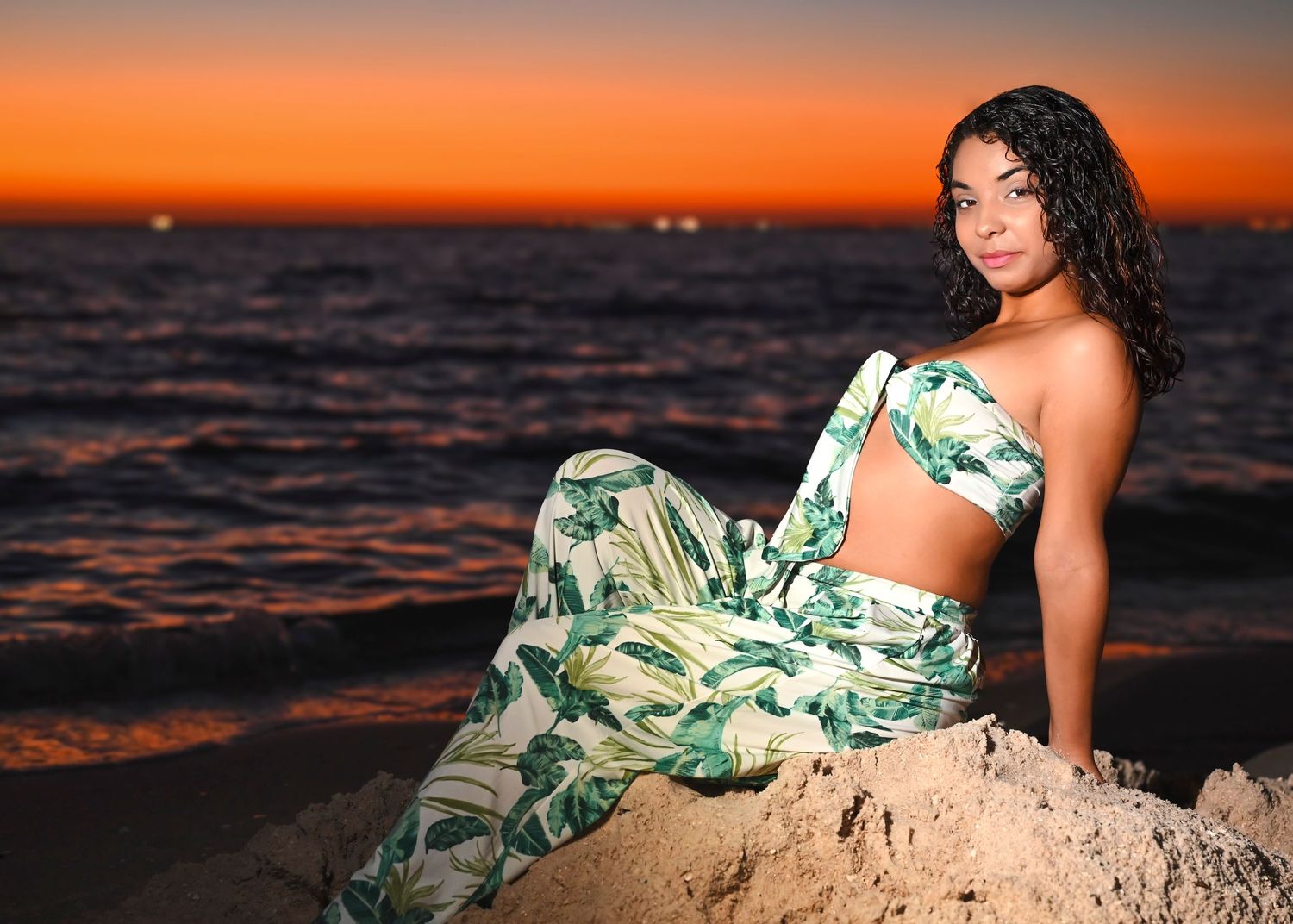 Woman in floral two-piece outfit poses on rocks at a stunning orange sunset beach.