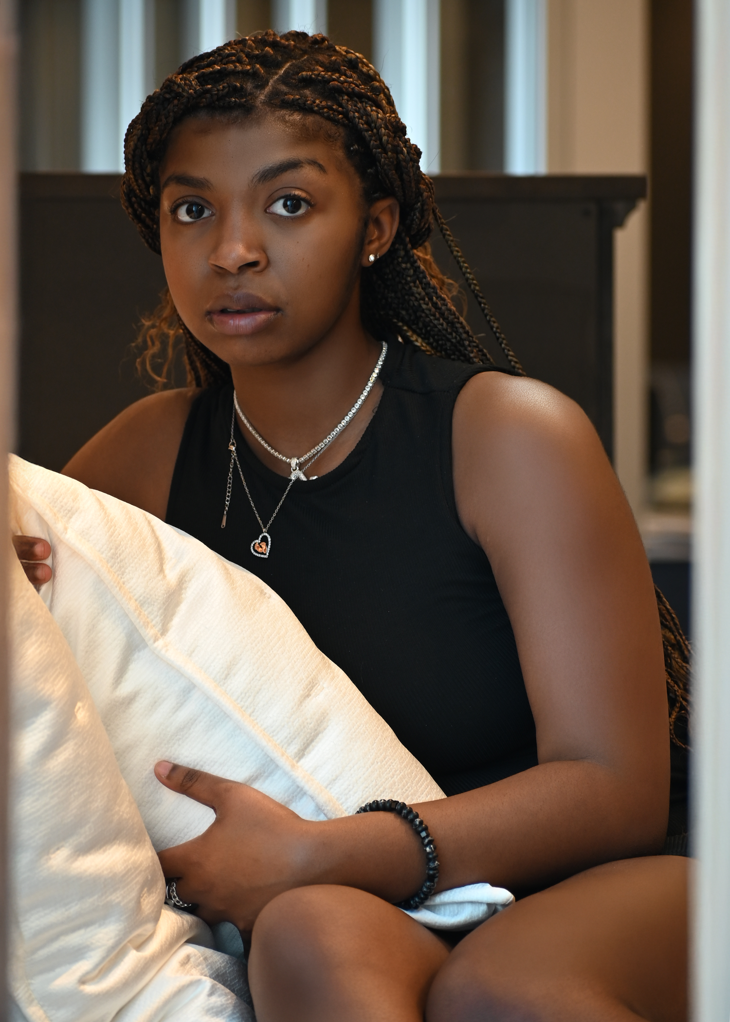 Indoor portrait wearing black sleeveless top with braided hair and layered necklaces.