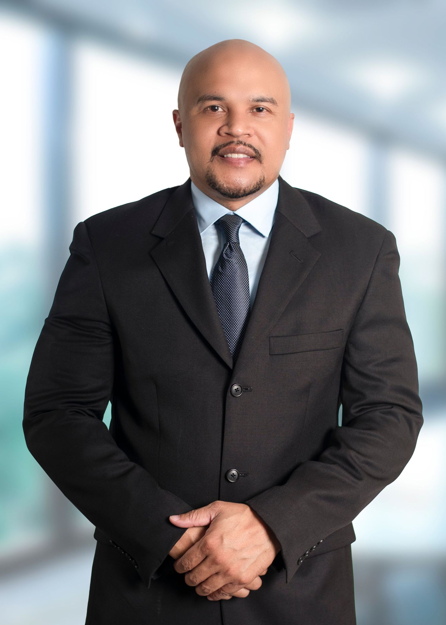Bald man with a goatee in a dark business suit, light blue shirt and gray tie, smiling with hands clasped in a bright modern office.