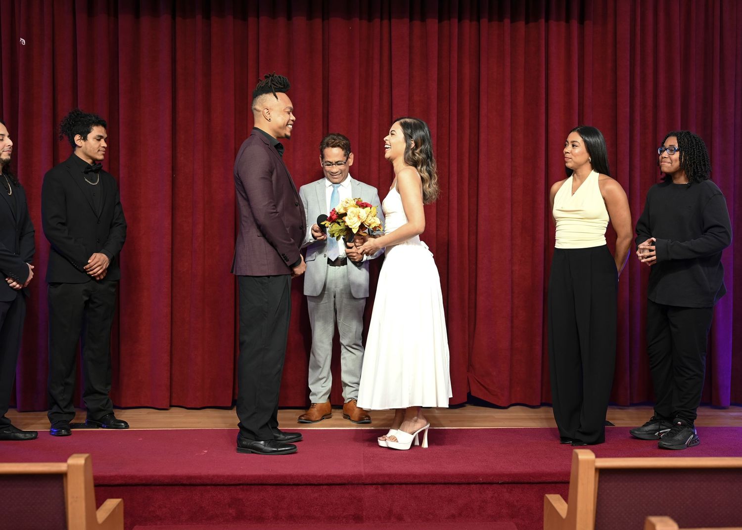 Couple exchanging vows at a wedding ceremony on stage with guests standing nearby, officiant in gray suit.