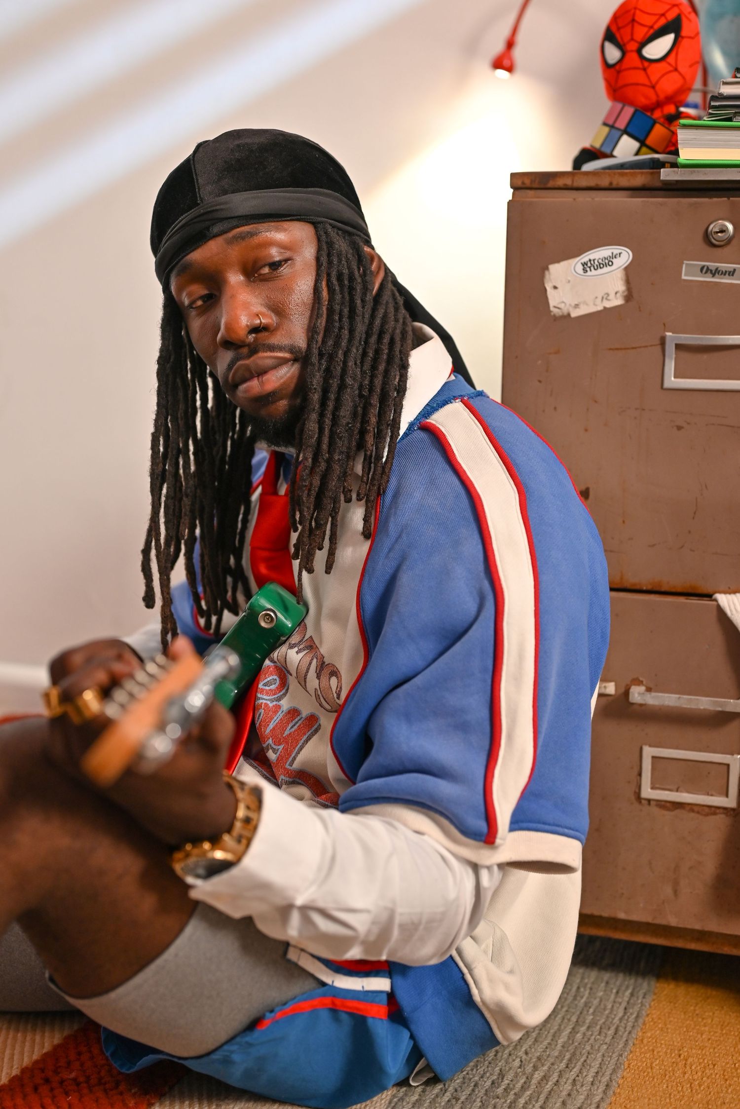 Person with dreadlocks playing guitar while sitting on floor near filing cabinet.