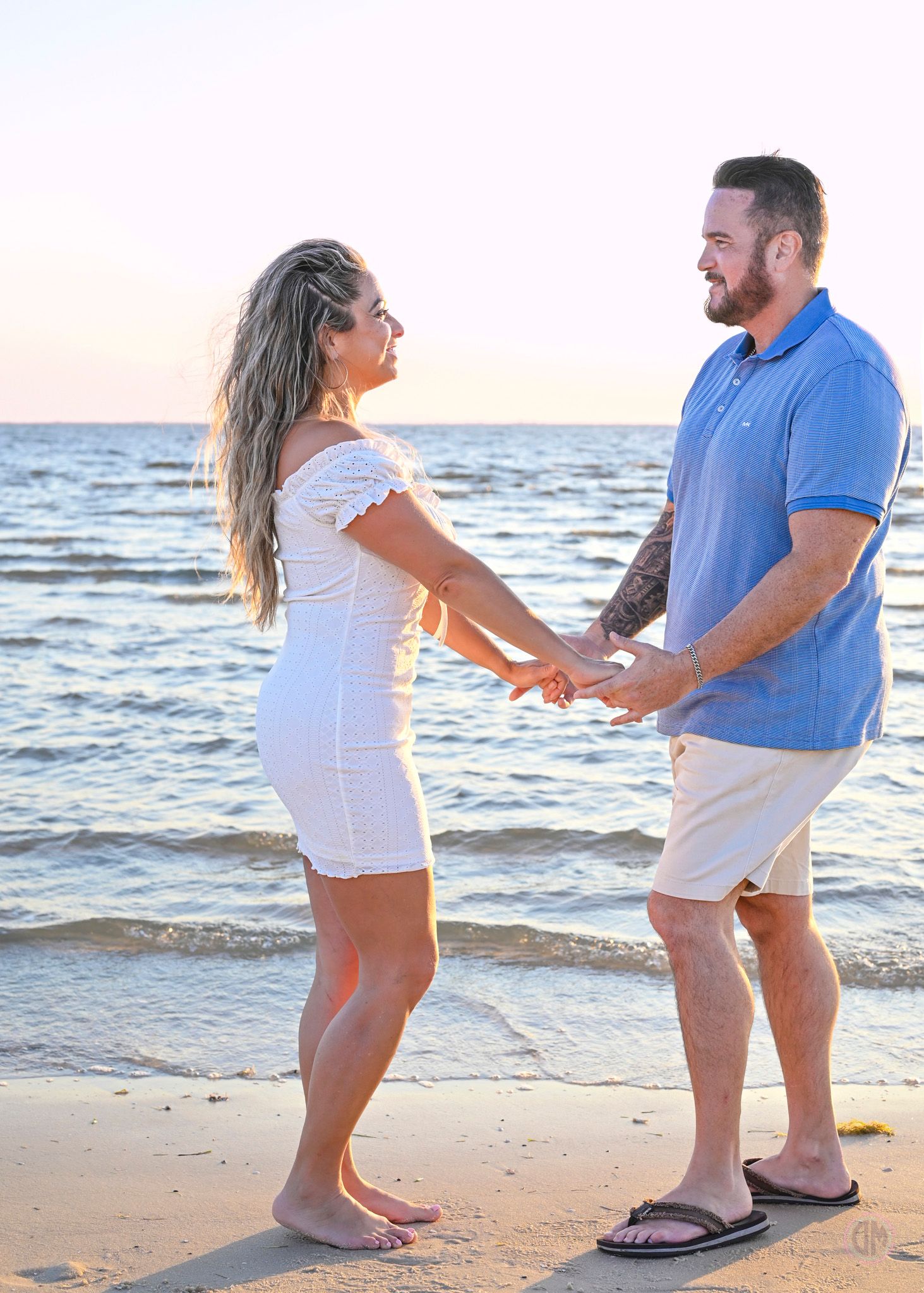 Couple holding hands on a sandy beach at sunset, woman in white dress and man in blue polo shirt.