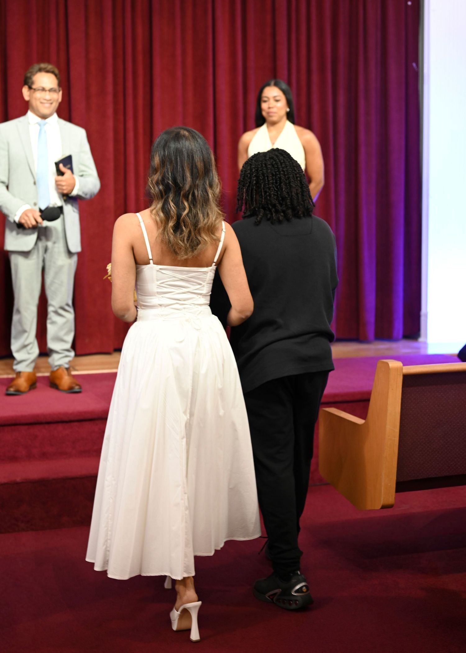 Bride in a white dress and a partner in black face an officiant holding a book during a wedding ceremony in a chapel with red curtains.