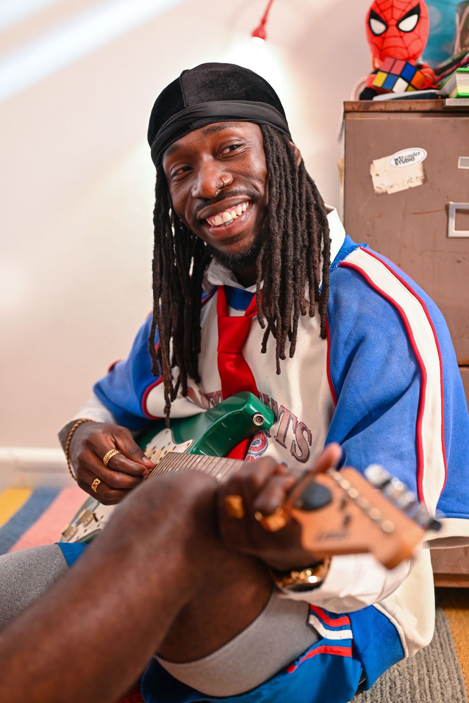 Person with dreadlocks smiling while playing guitar with Spider-Man decoration visible.
