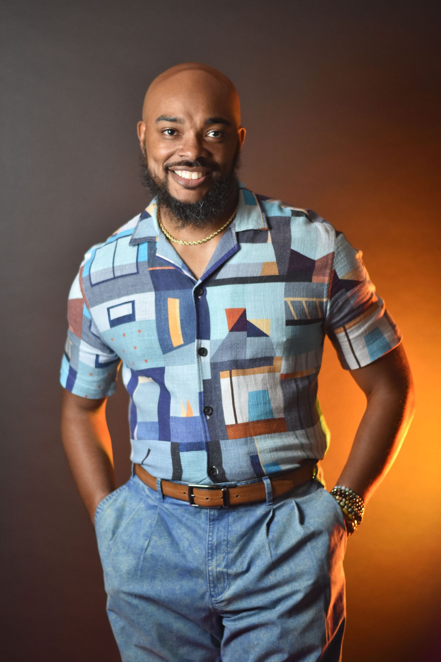 Professional studio portrait showing a stylish geometric patterned short-sleeve button-up shirt and dress pants against orange backdrop.