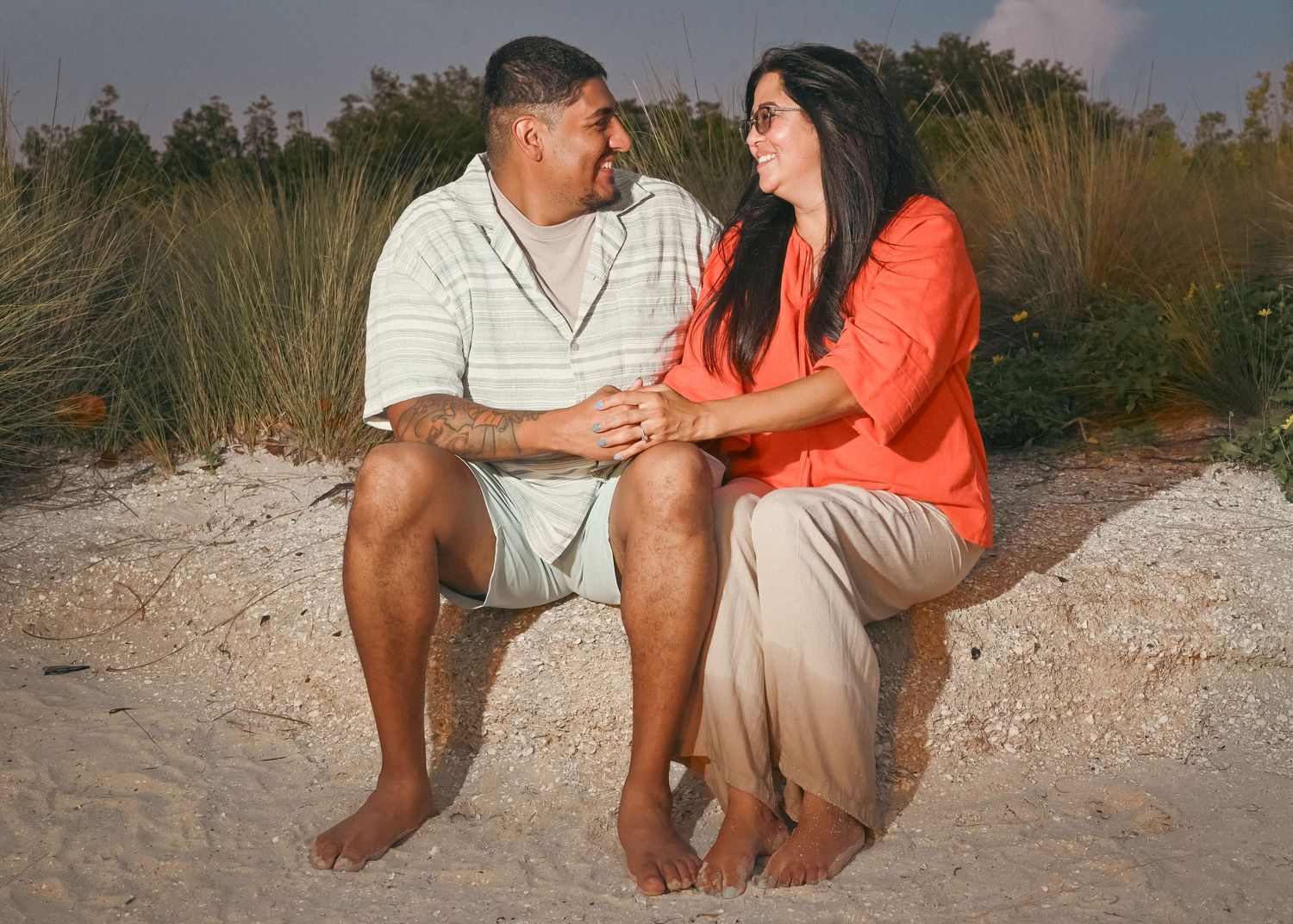 Couple sitting together on sandy beach, holding hands and smiling, with beach grass in background.