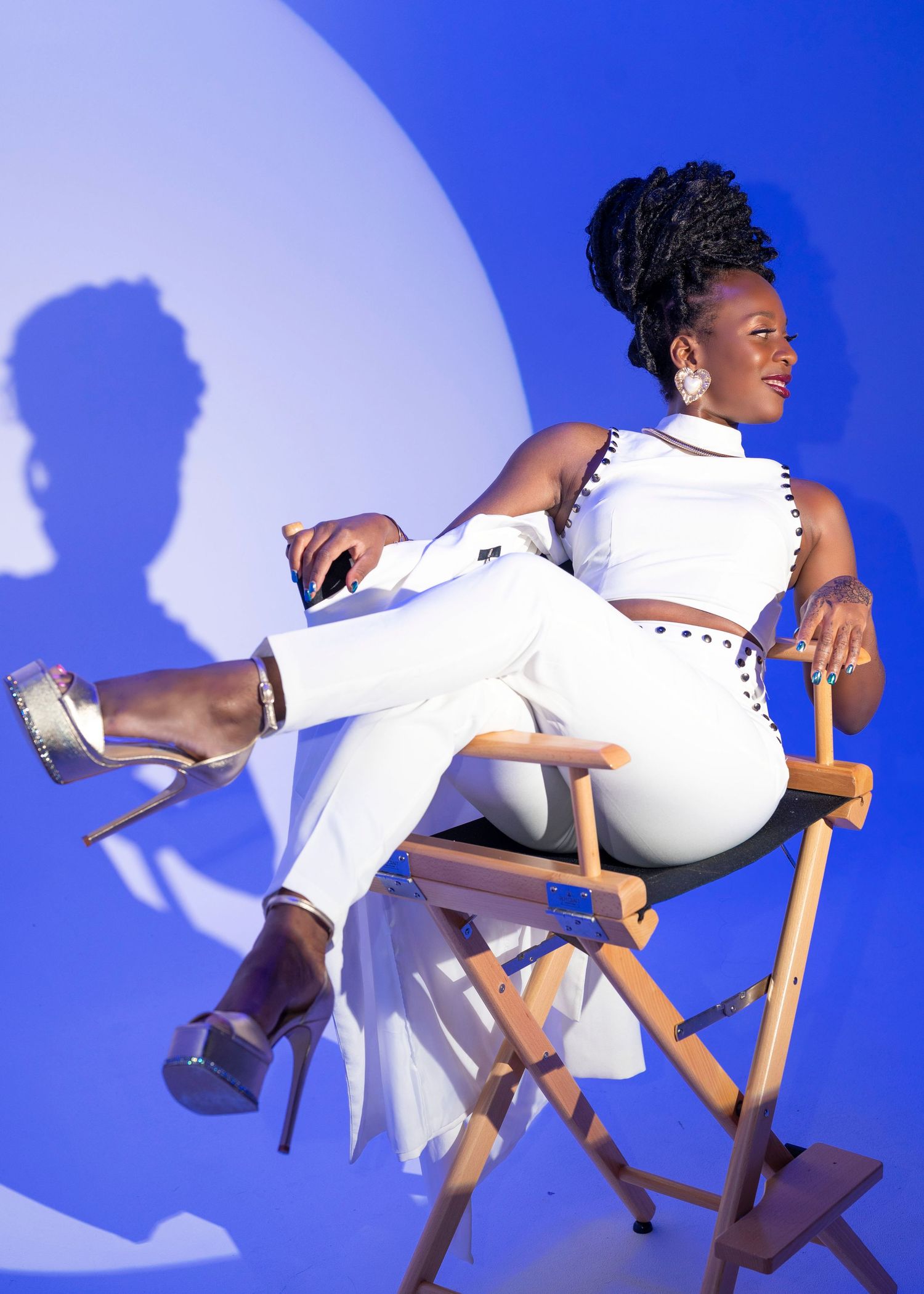 Model in white studded jumpsuit poses dramatically on director's chair against blue backdrop.