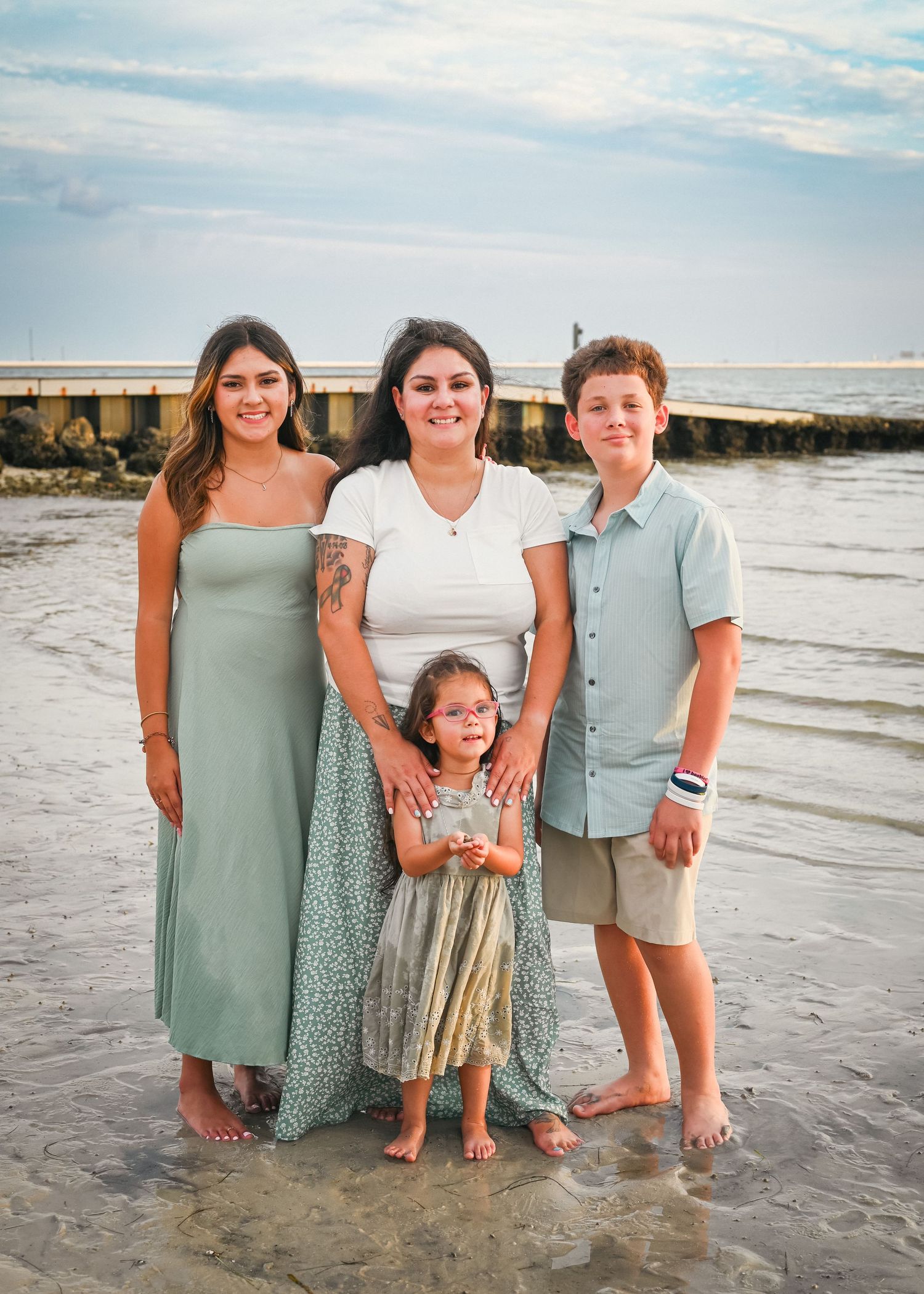 Family of four smiling together on a beach, mother with two teens and a toddler, wearing summer outfits near the water.