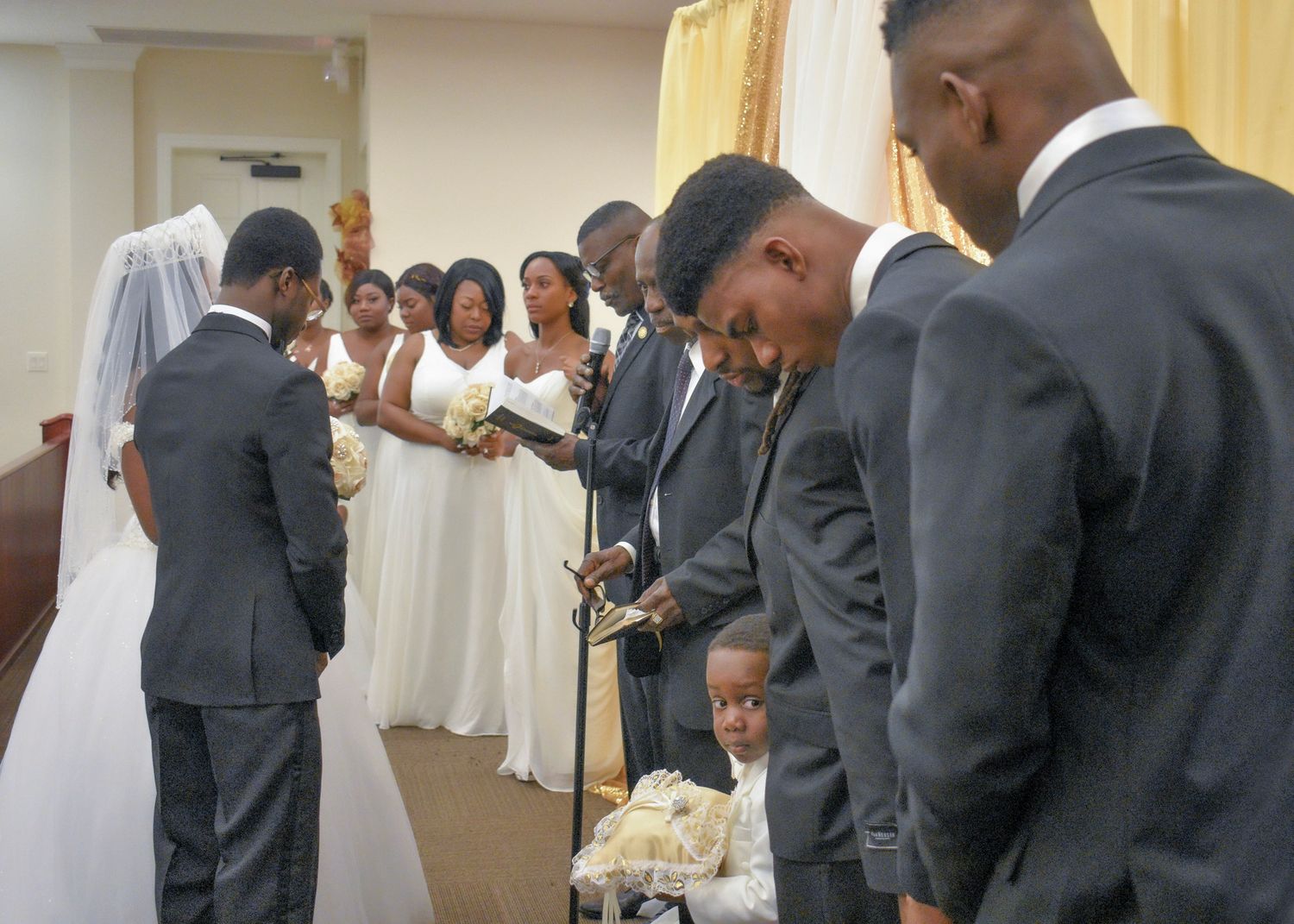 Wedding ceremony with bridal party bowing in prayer, bride in white gown, groomsmen in gray suits inside a church.