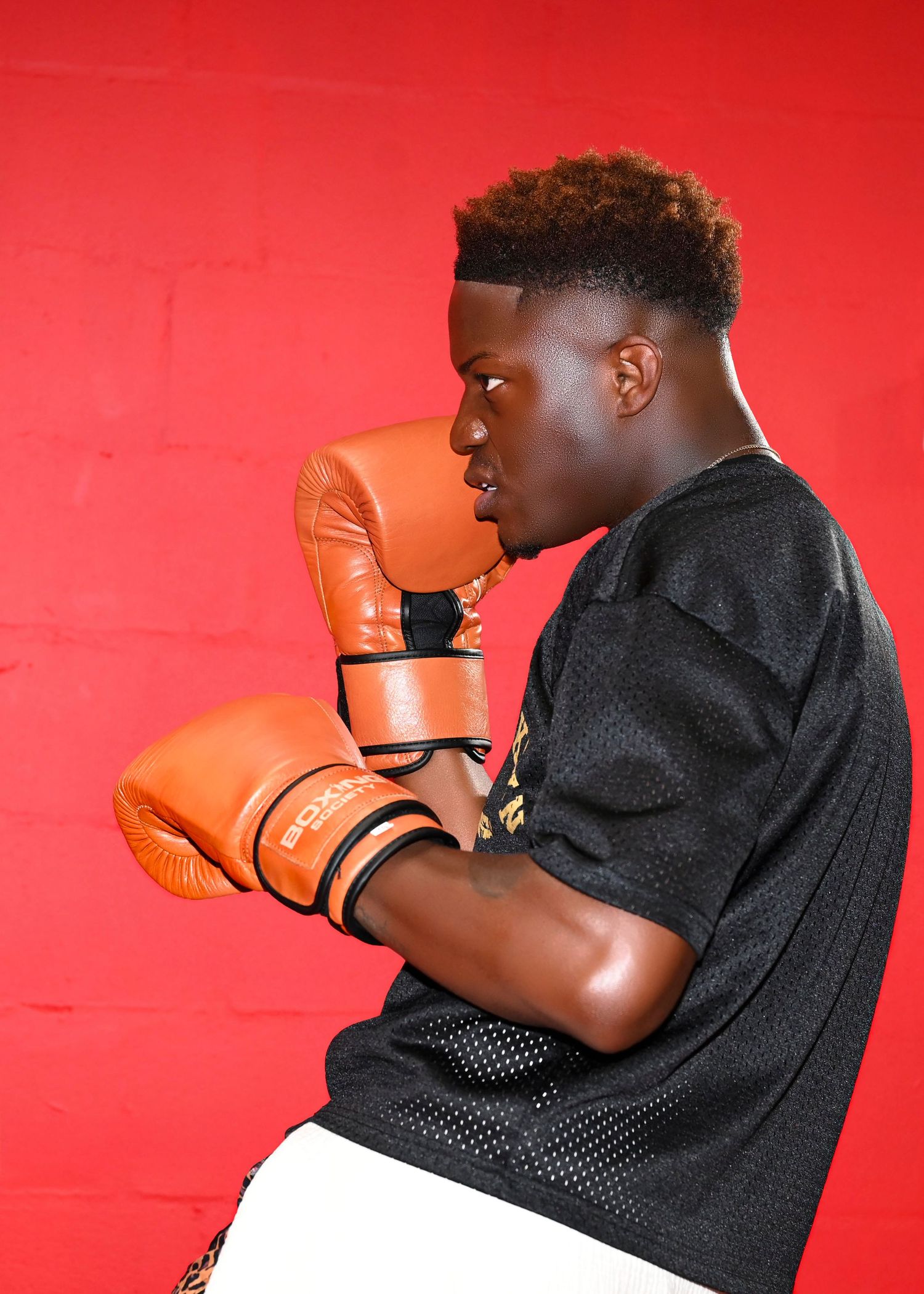 Boxer in black mesh jersey and orange gloves poses in fighting stance against red background.