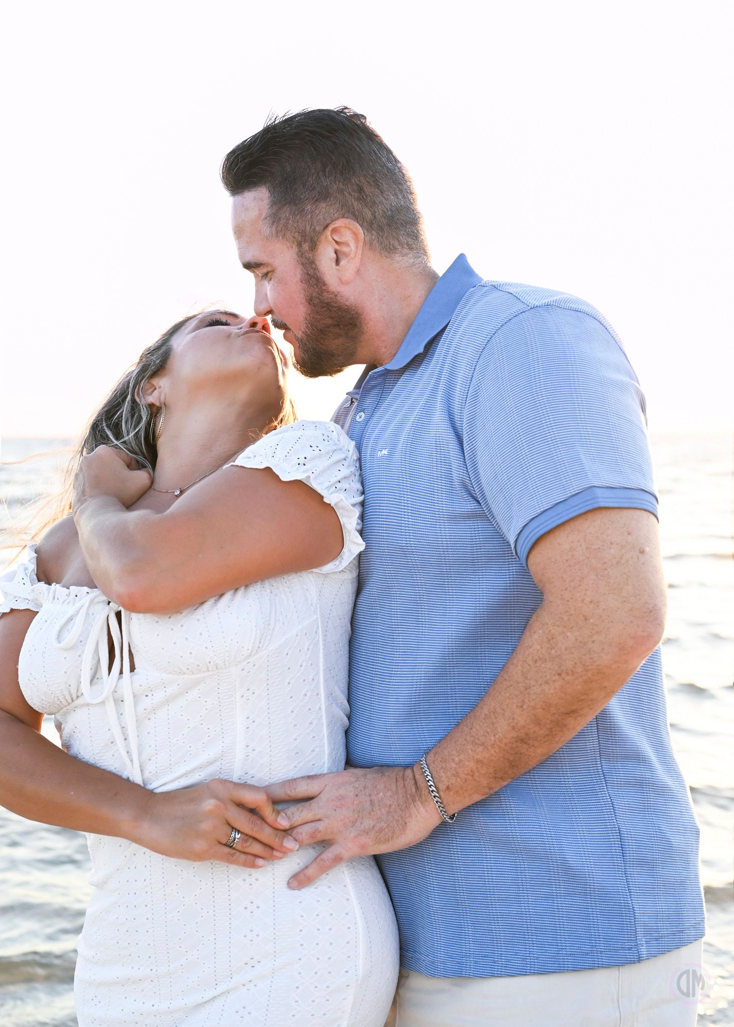 Romantic couple embracing and kissing on beach, woman in white dress and man in blue polo shirt.